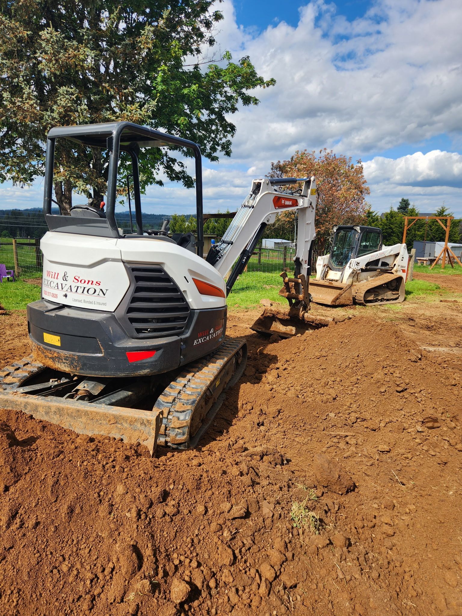 Small white excavator on a dirt lot with a Bobcat skid steer in the background. Overcast sky.