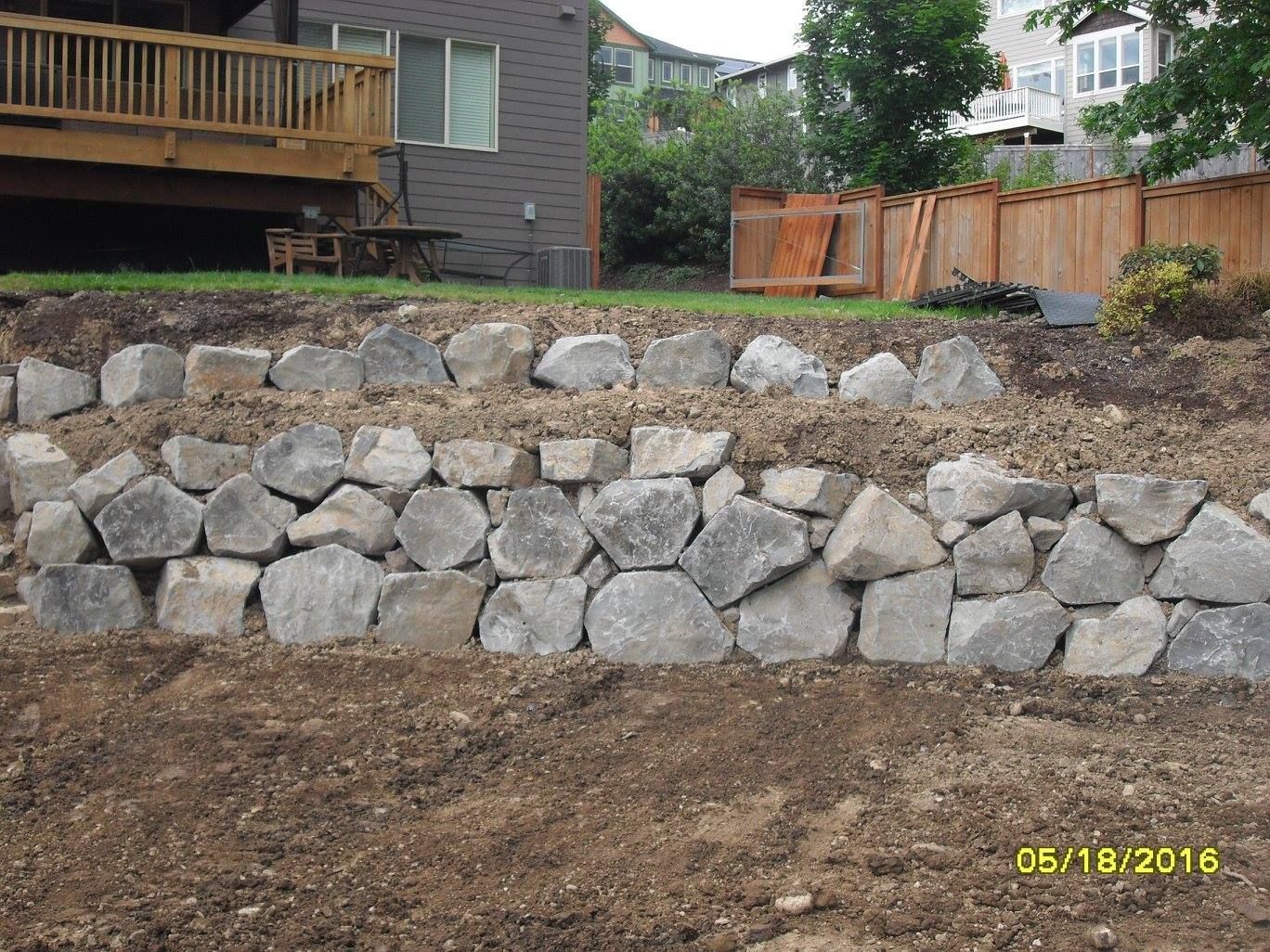 Rock retaining wall on a hillside in a backyard, brown soil in foreground, deck and house in background.