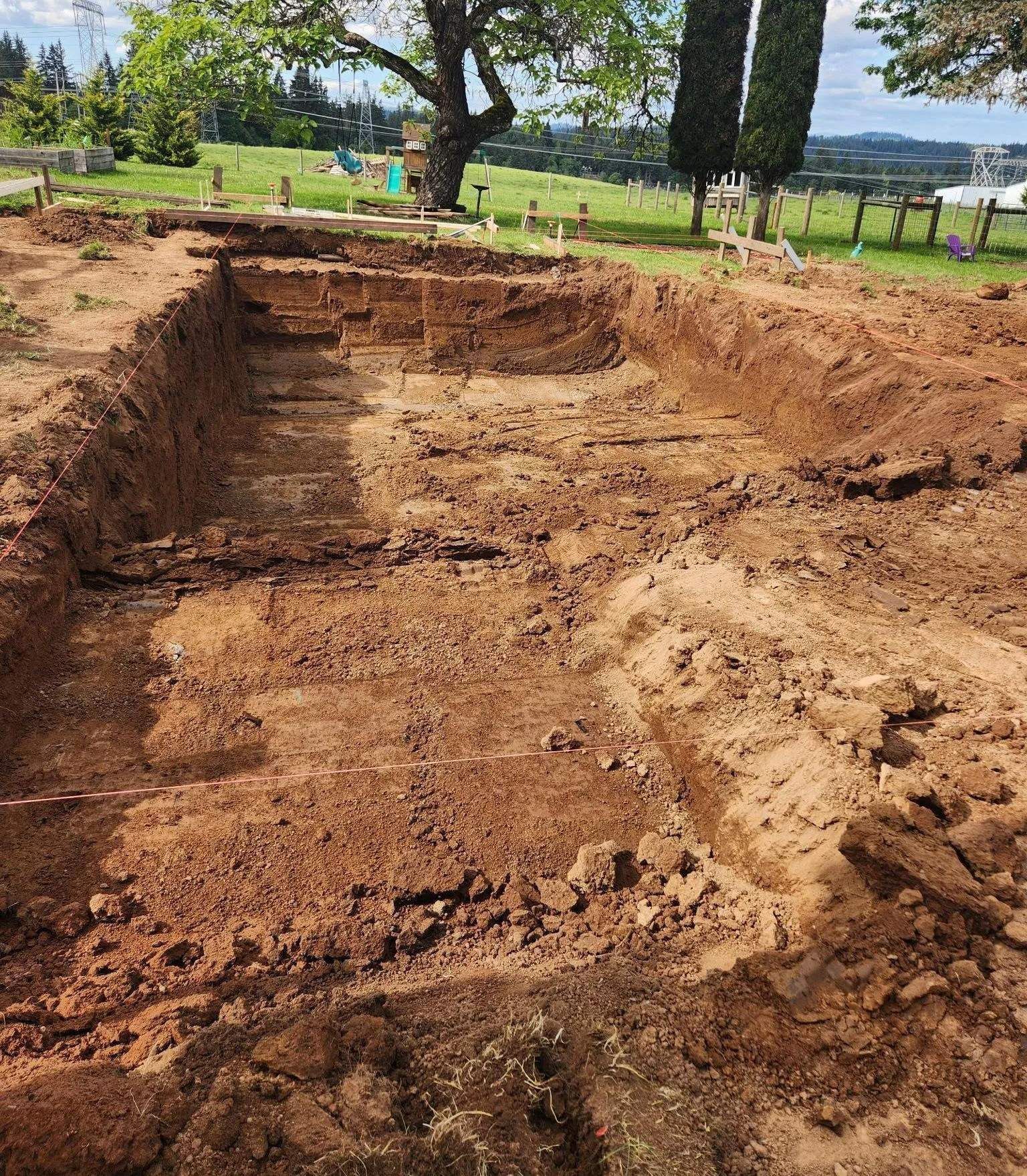 Excavated rectangular area in brown dirt with surrounding grassy landscape and trees in the background.
