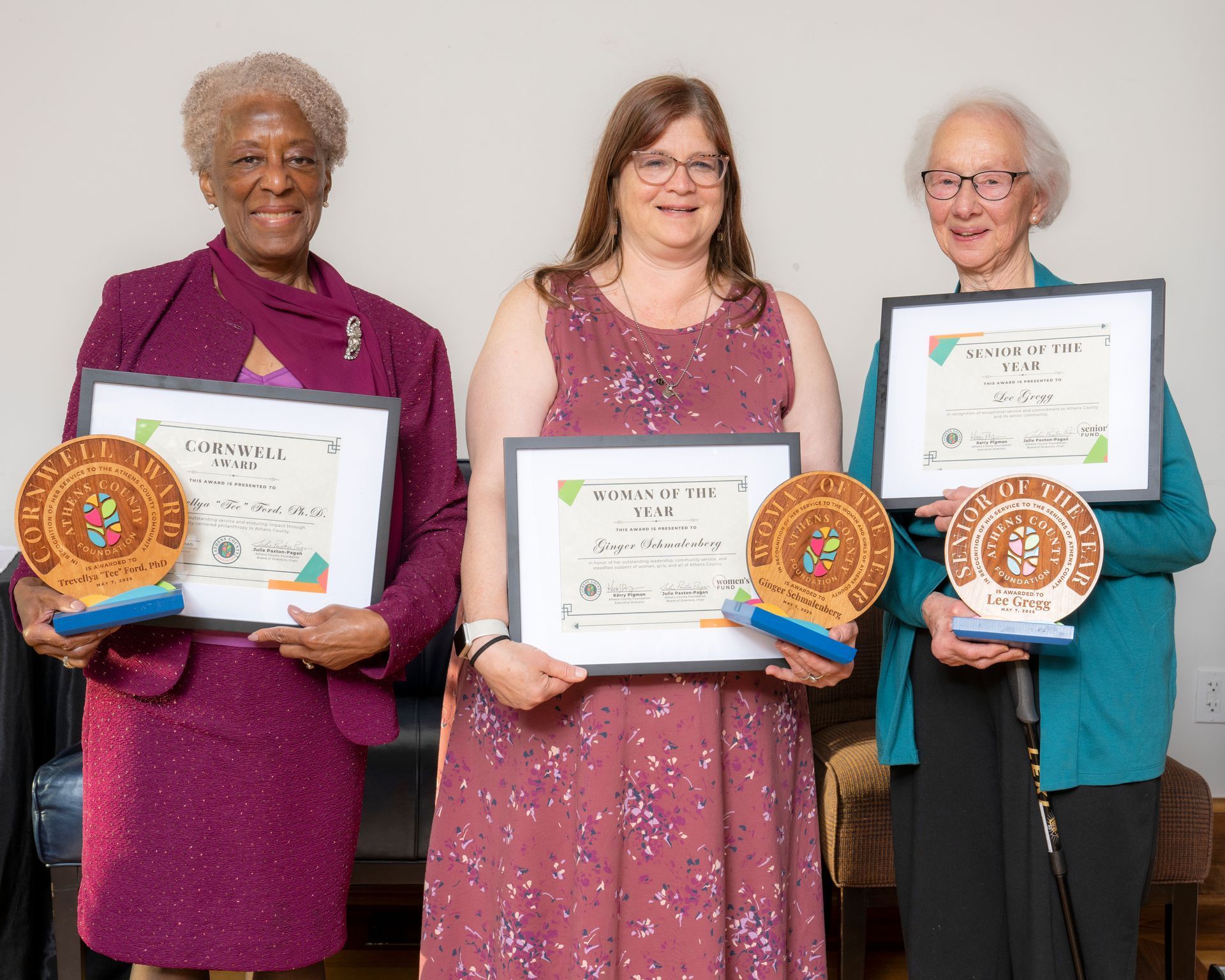 Three women standing together, smiling, and holding framed certificates and wooden awards. 