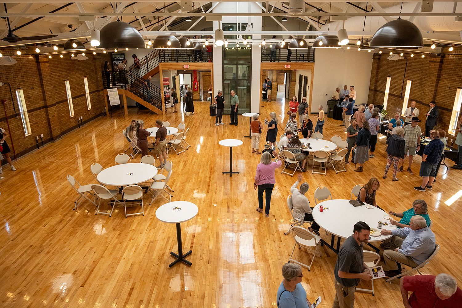 People mingling in a large event space with round tables and a wood floor. Athens Armory Community Open House