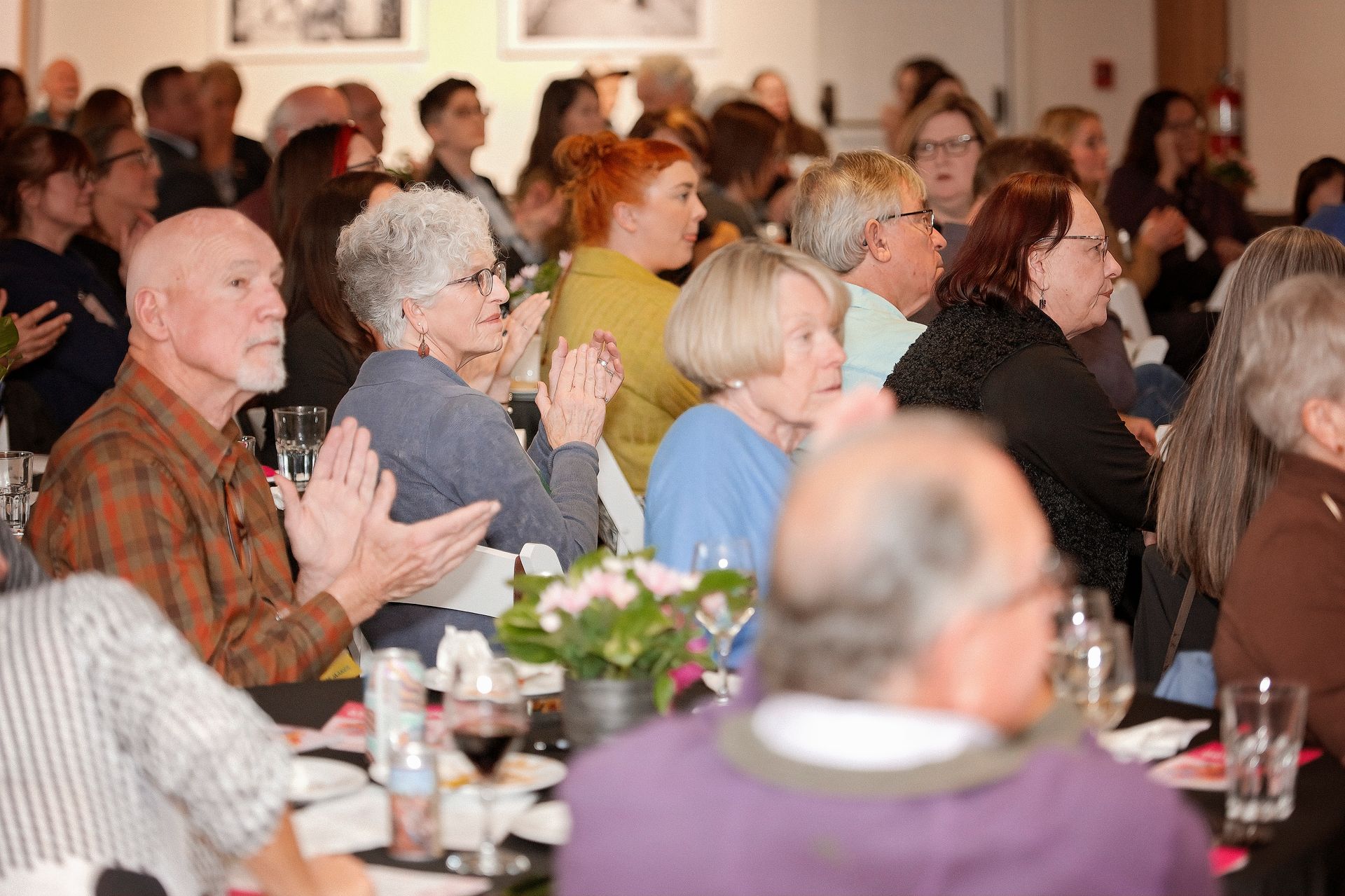 A large group of people are sitting at tables in a room applauding.