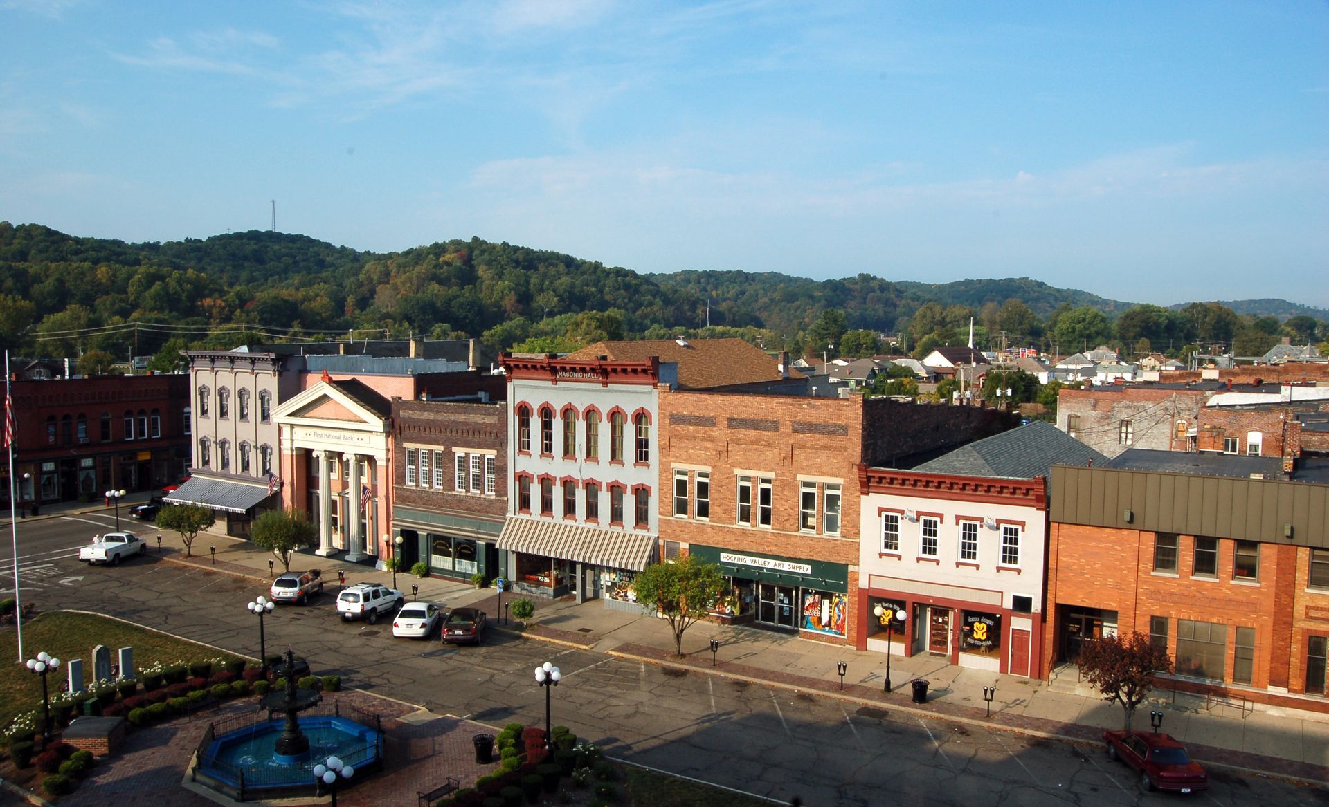 Row of brick buildings with shops and cars parked on a town square, green hills in the background under a blue sky.