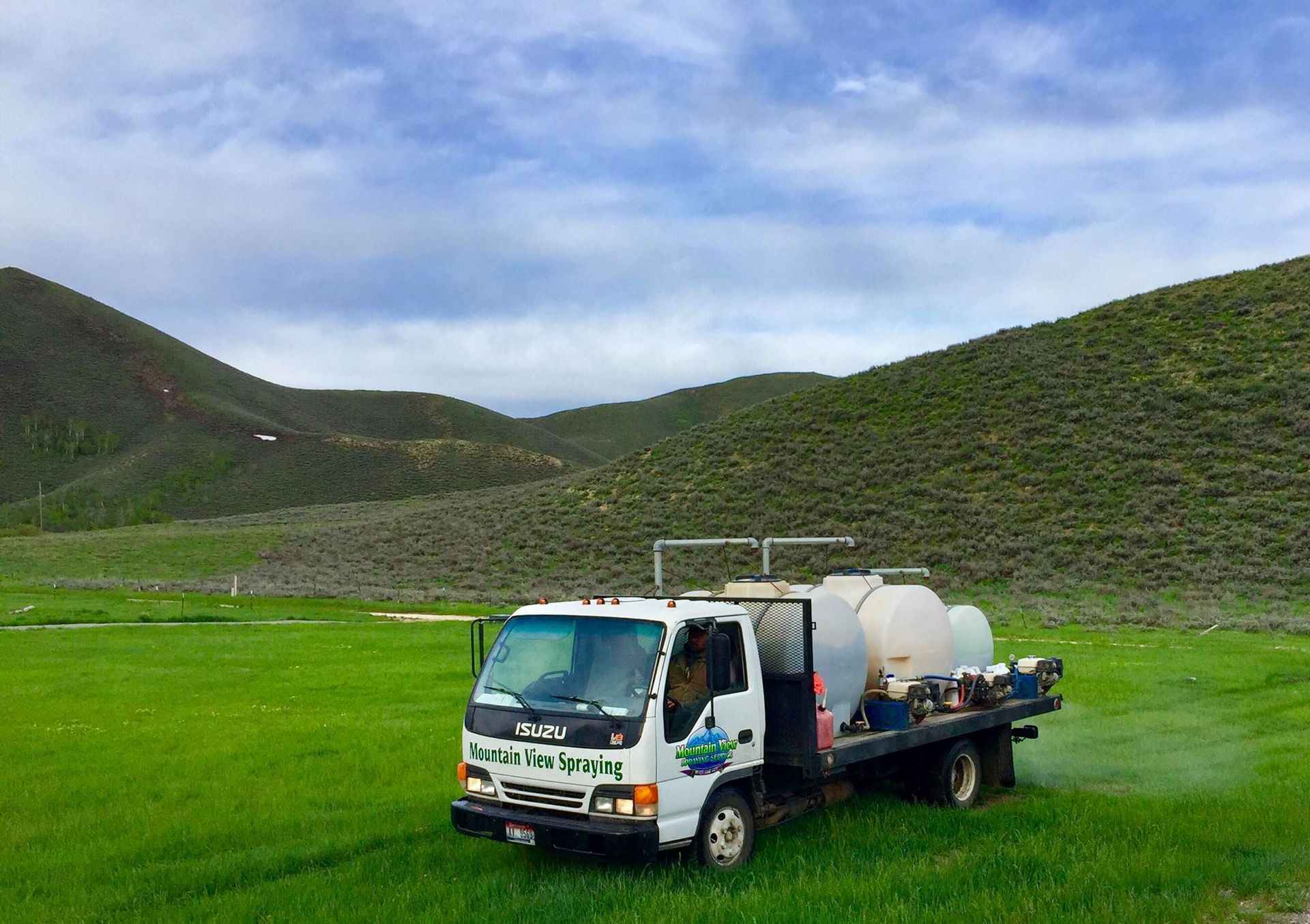 A white flatbed truck with large water tanks parked in a lush green field beneath rolling, grass-covered hills.