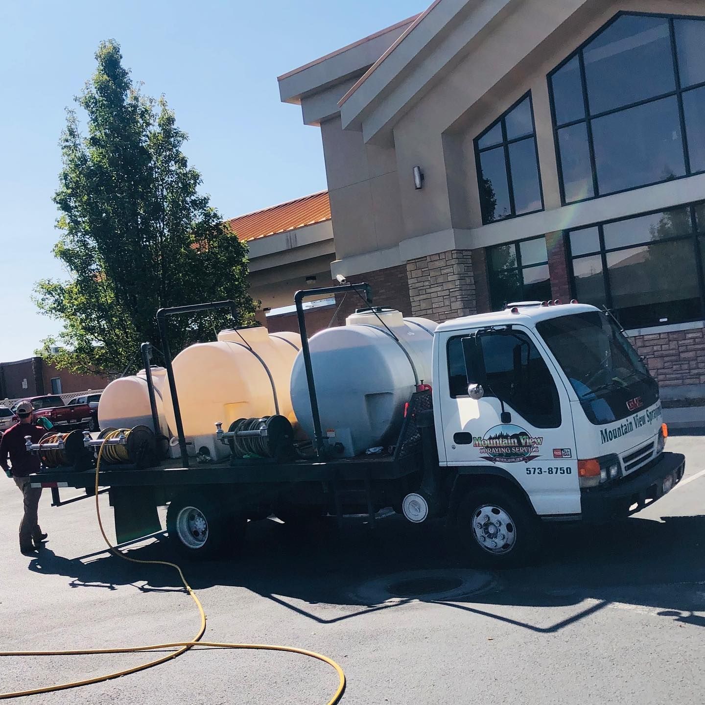 A white utility truck with three large water tanks mounted on its flatbed parked in front of a modern brick building.