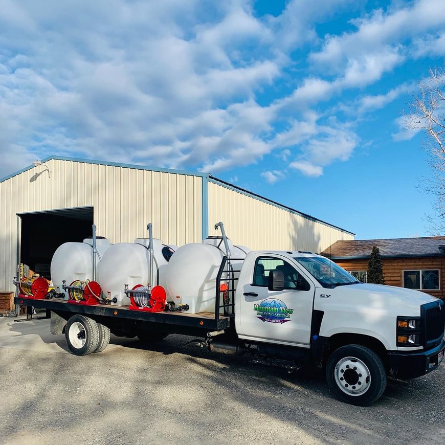 A white flatbed truck parked in front of a metal building, carrying three large white liquid storage tanks.