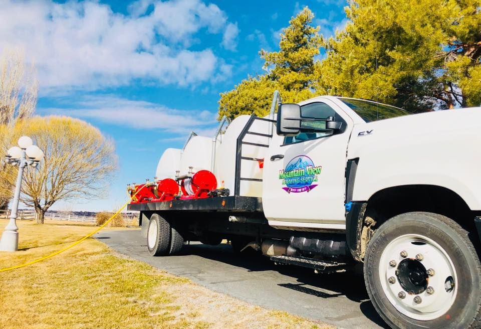 A white service truck carrying large water tanks parked on a paved road near trees under a bright blue sky.