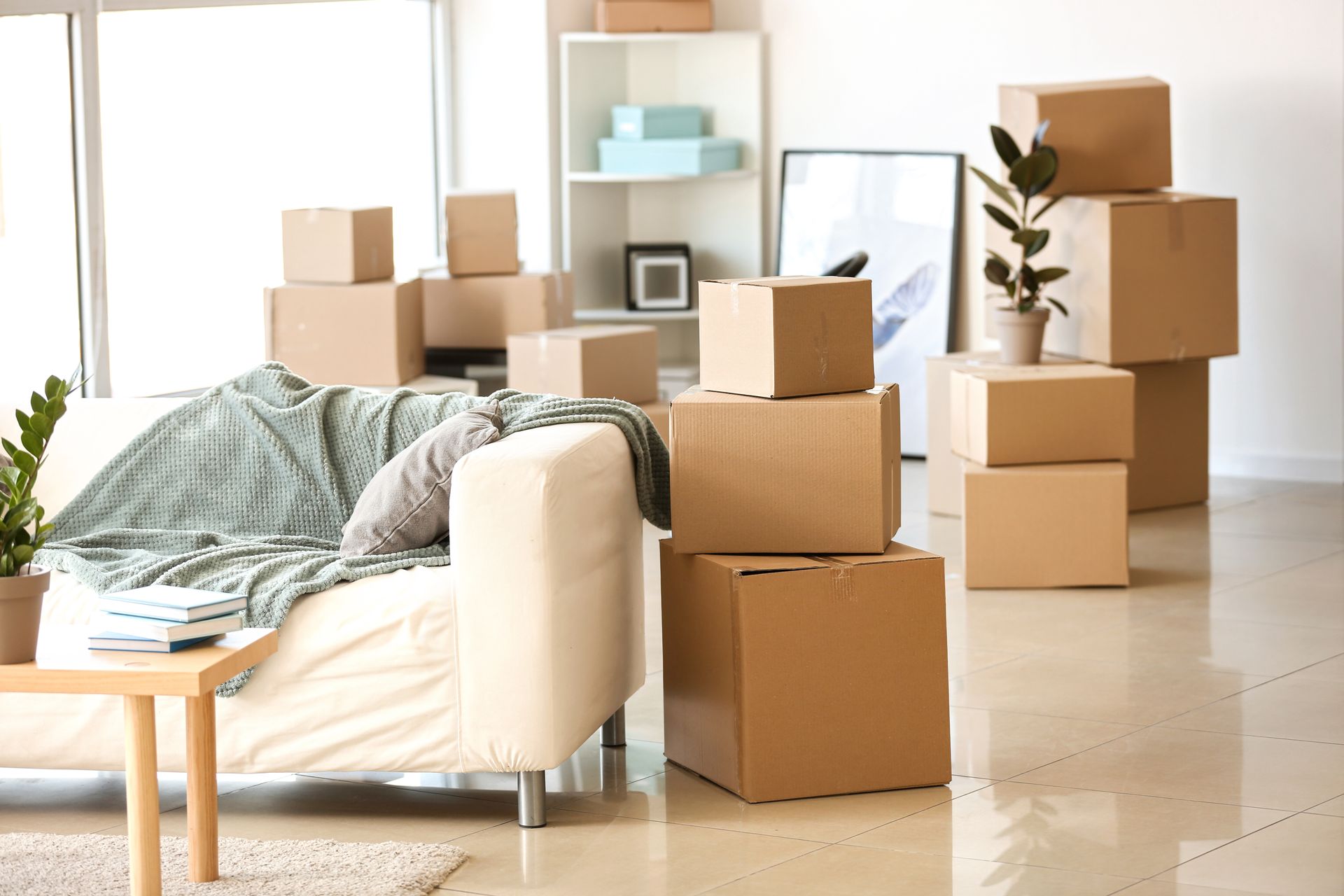 Living room with stacked moving boxes, a couch, and unpacked household items in a bright apartment