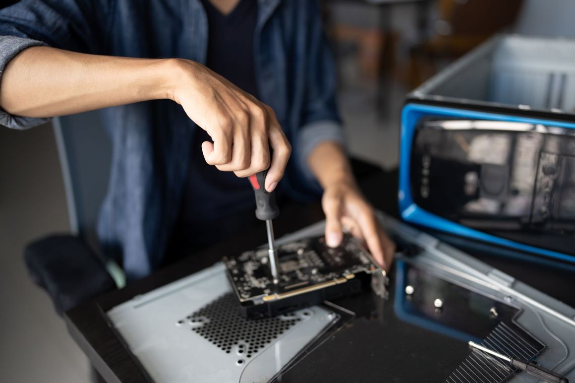 Hands using a screwdriver to repair a computer motherboard beside a blue microscope-like device