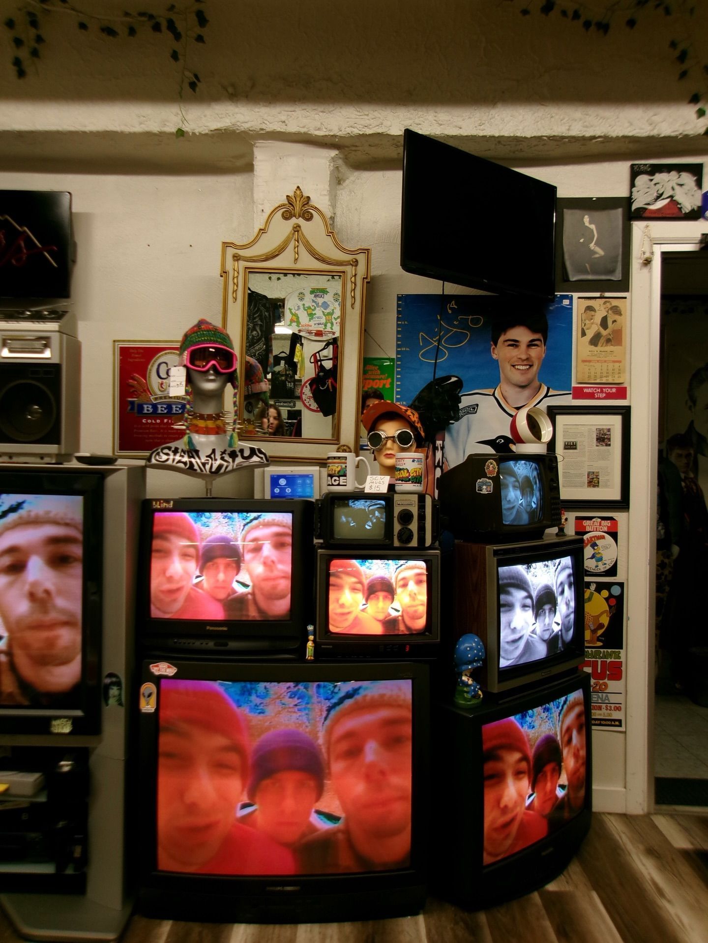 Wall of multiple TVs showing a red-tinted face, with a mirror, framed photos, and small decor items nearby