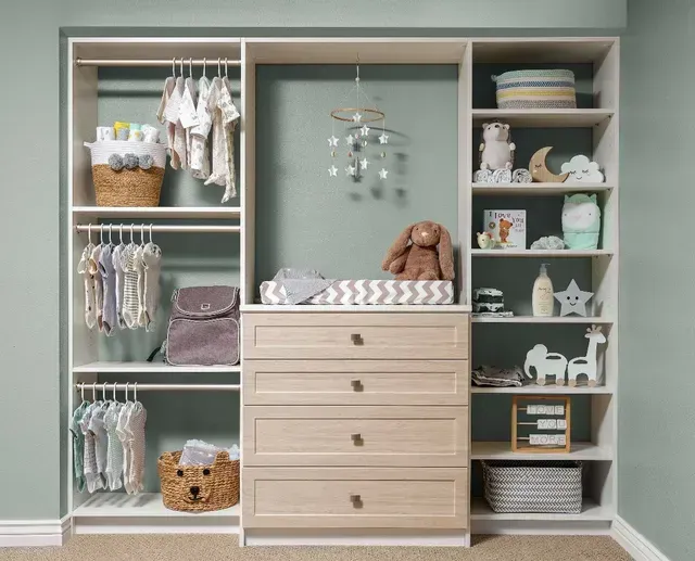 A white nursery closet with shelving, drawers, and hanging clothes; a changing pad and toys are visible.