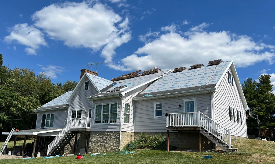 A two-story house undergoing roof replacement, with roofing materials and underlayment visible on the roof.
