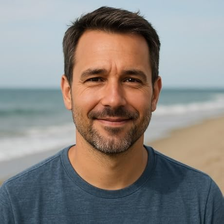 Man smiling at the beach, wearing a blue shirt with ocean and sandy shore behind him