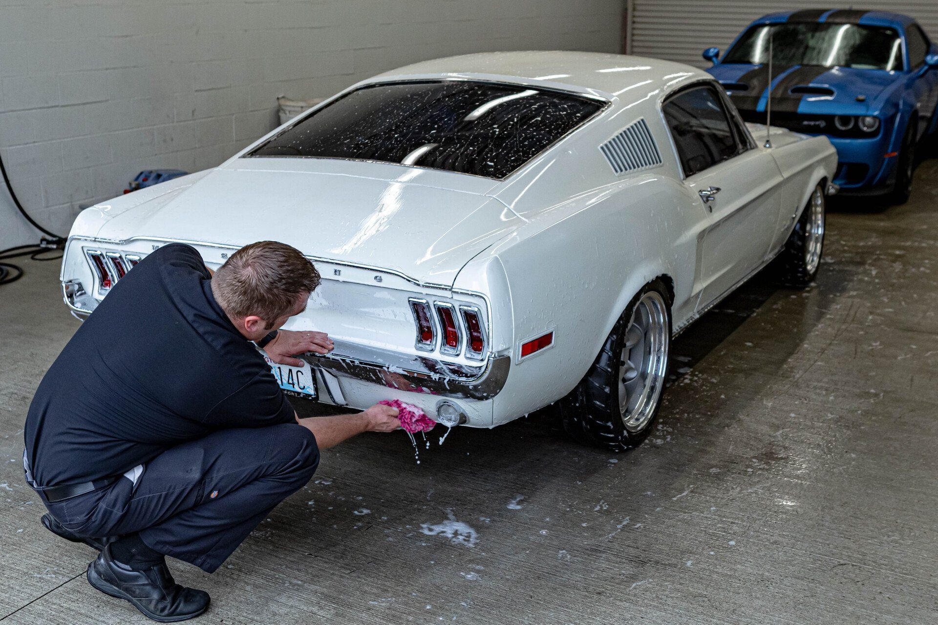 A man is kneeling down to wash a white mustang in a garage.
