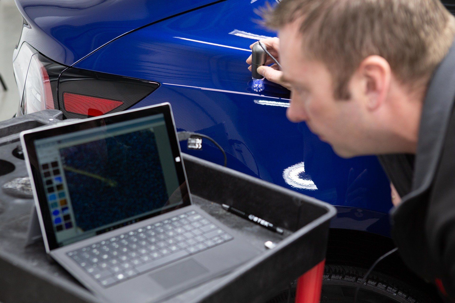 A man is looking at a blue car while using a laptop