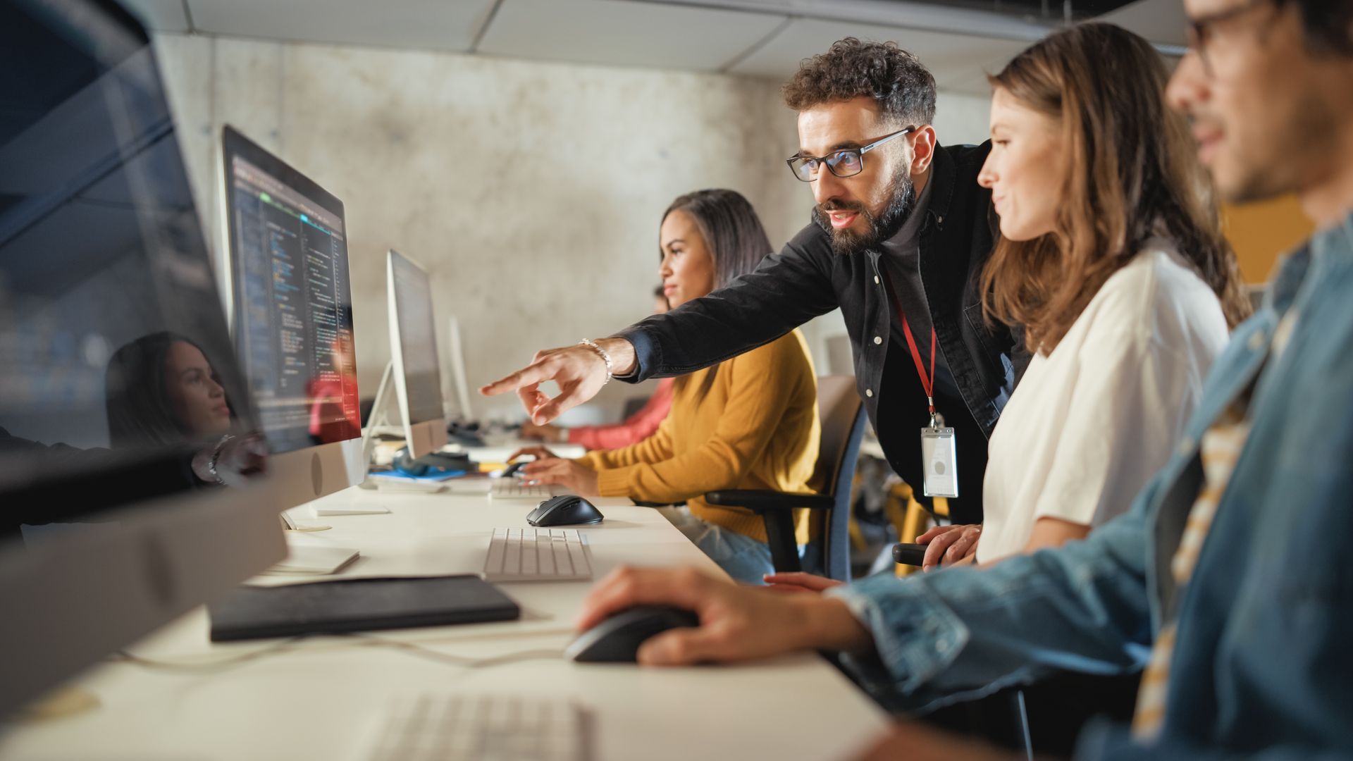 Man points at a computer screen, assisting two colleagues in a brightly lit office.