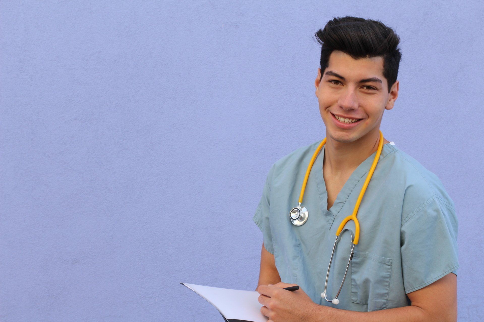 A BLS Provider, a young man wearing scrubs and a stethoscope is holding a clipboard and smiling .