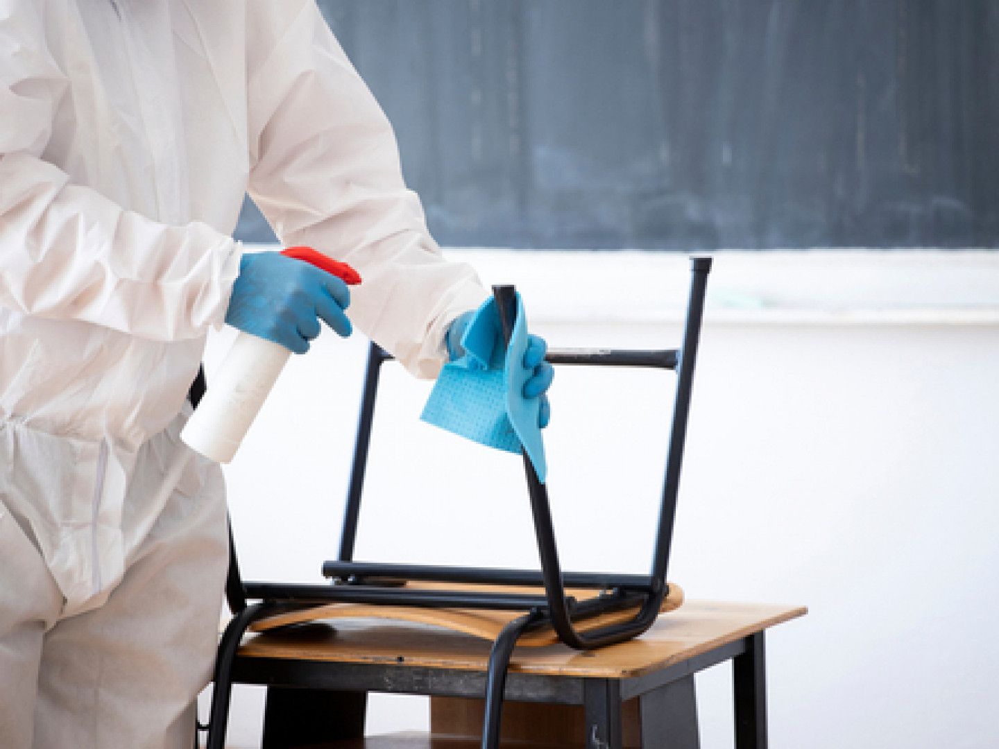 A person in a protective suit is cleaning a chair in a classroom.