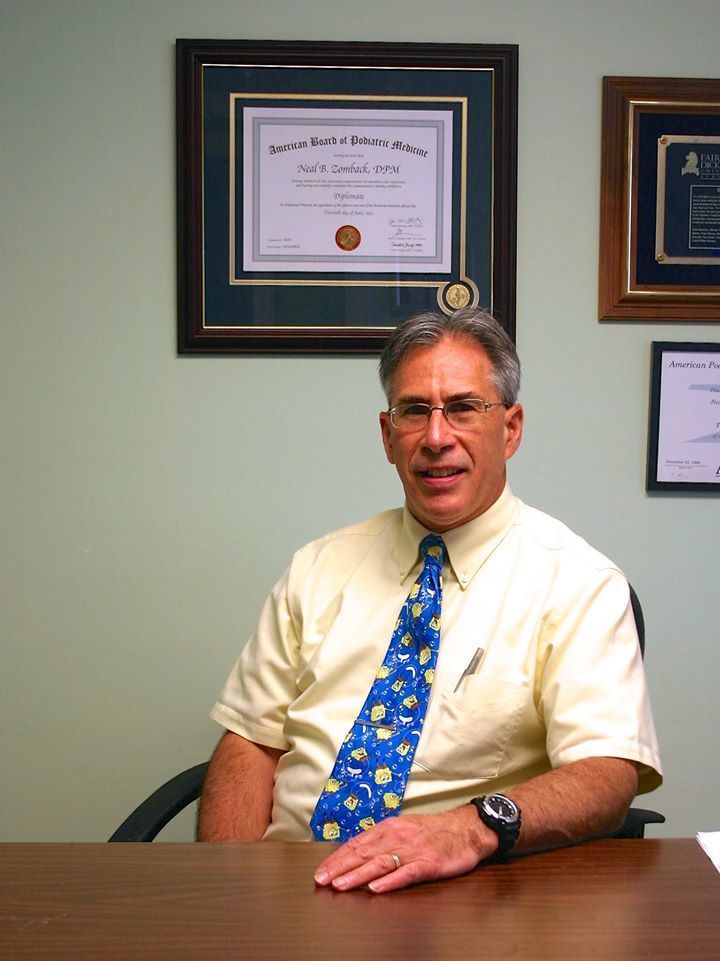 Man in a yellow shirt and blue tie sits at a desk in an office. Certificates hang on the wall.