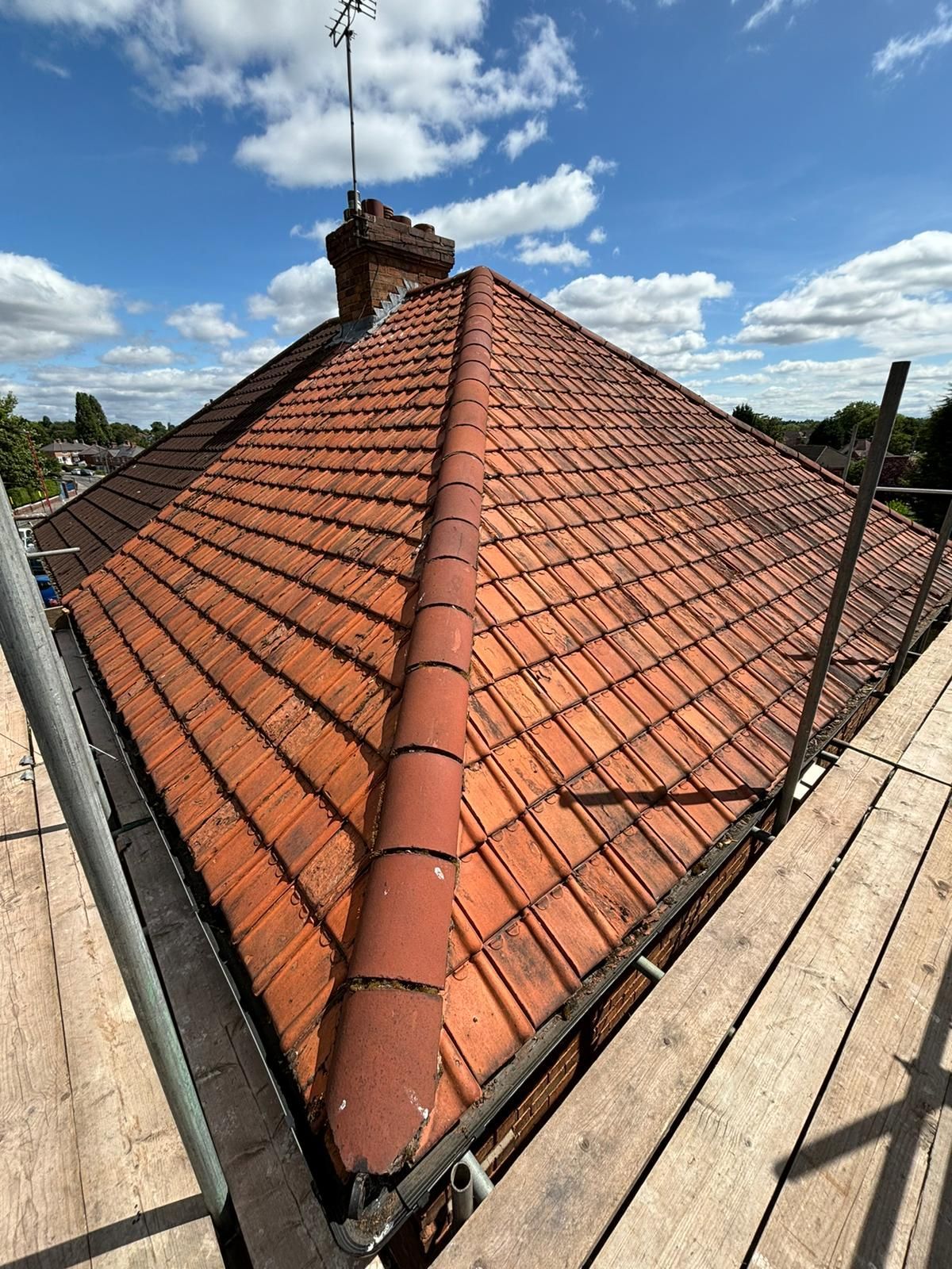 Red tiled roof with chimney, antenna, and scaffolding against a blue sky with clouds.