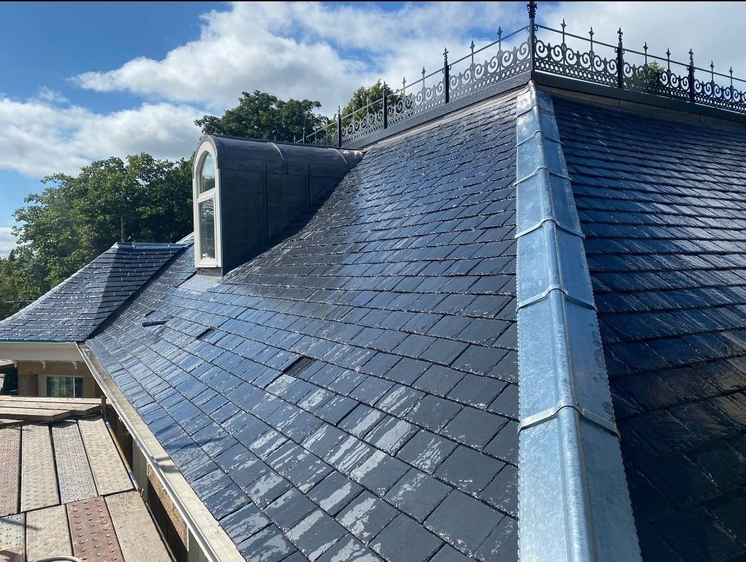 Slate roof with dormer window, metal trim, and decorative railing against a cloudy sky.
