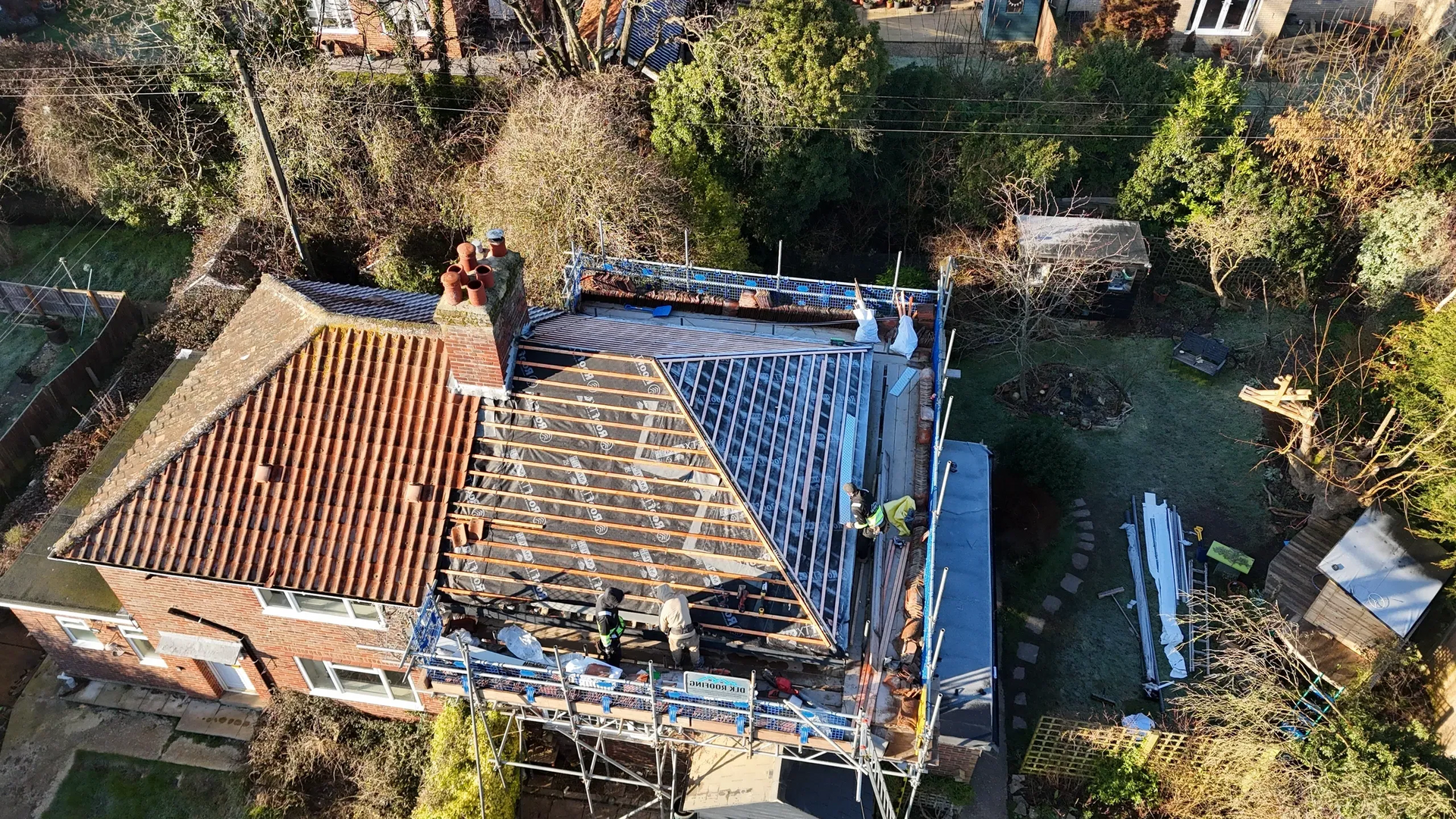 Aerial view of a house with scaffolding and roof tiles partially removed. Trees and yard surround the building.