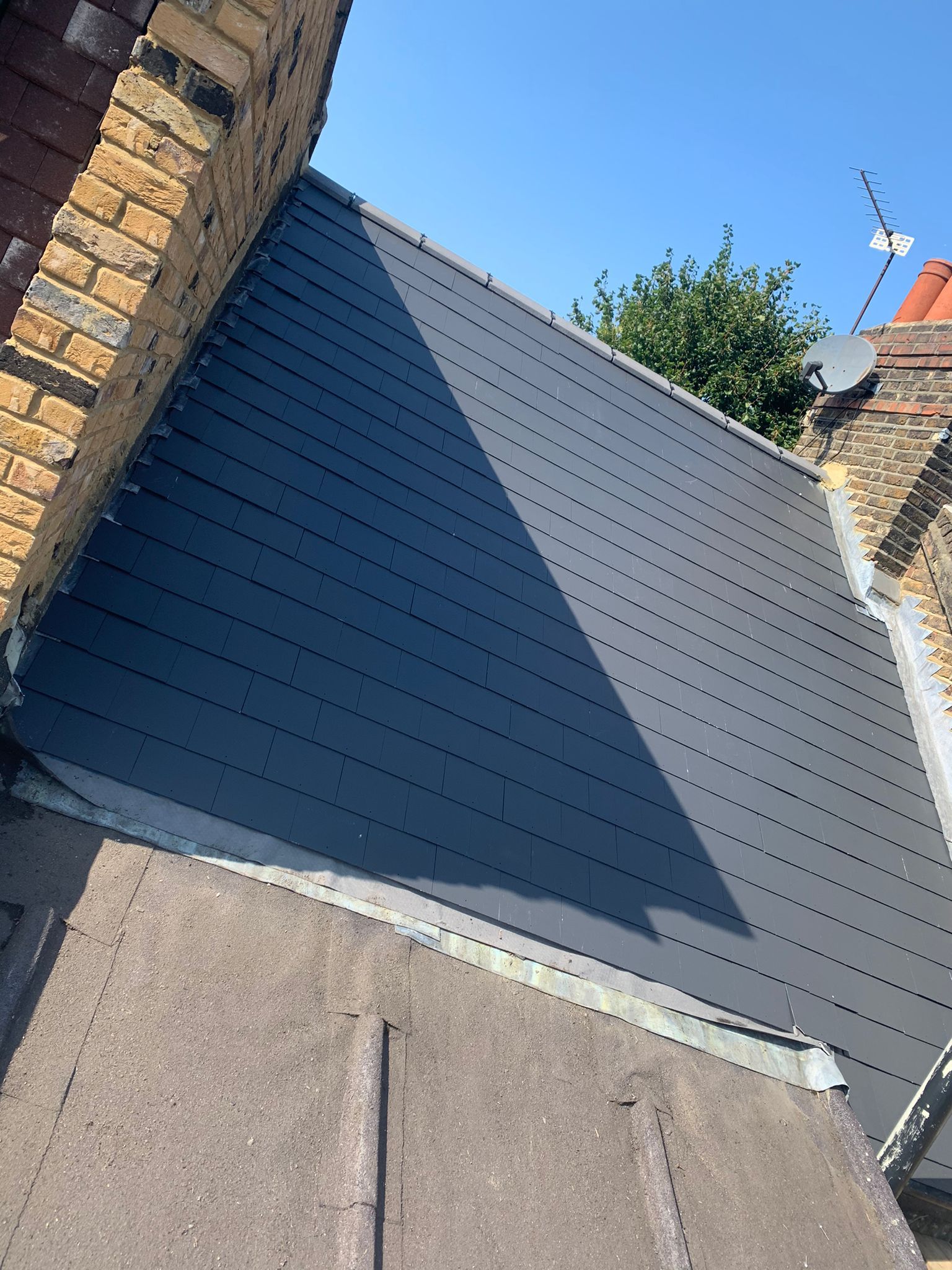 A section of a dark gray metal roof installed next to a brick chimney and other rooftops.