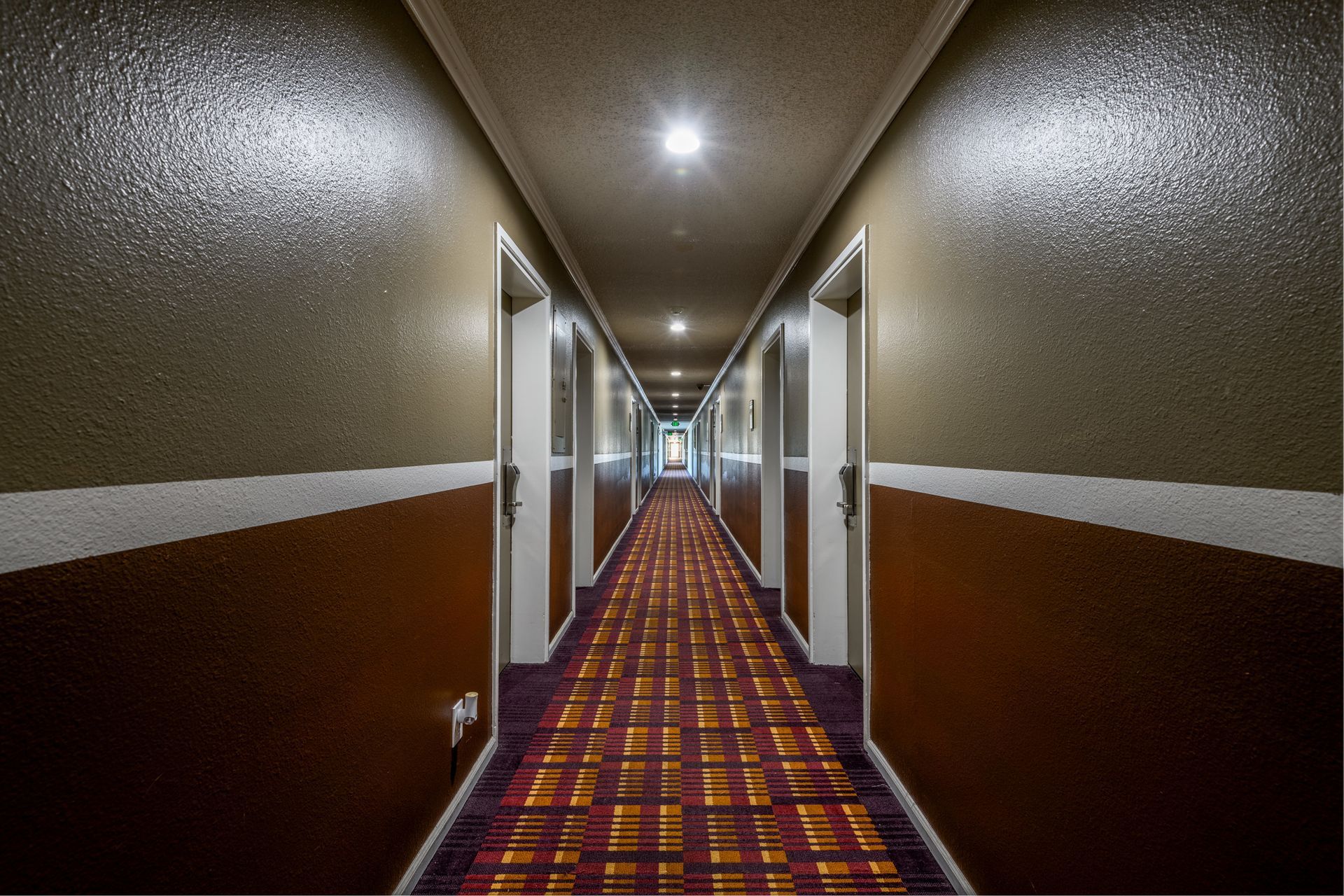 Hotel hallway with doors on both sides, a patterned red and gold carpet, and a light at the end.