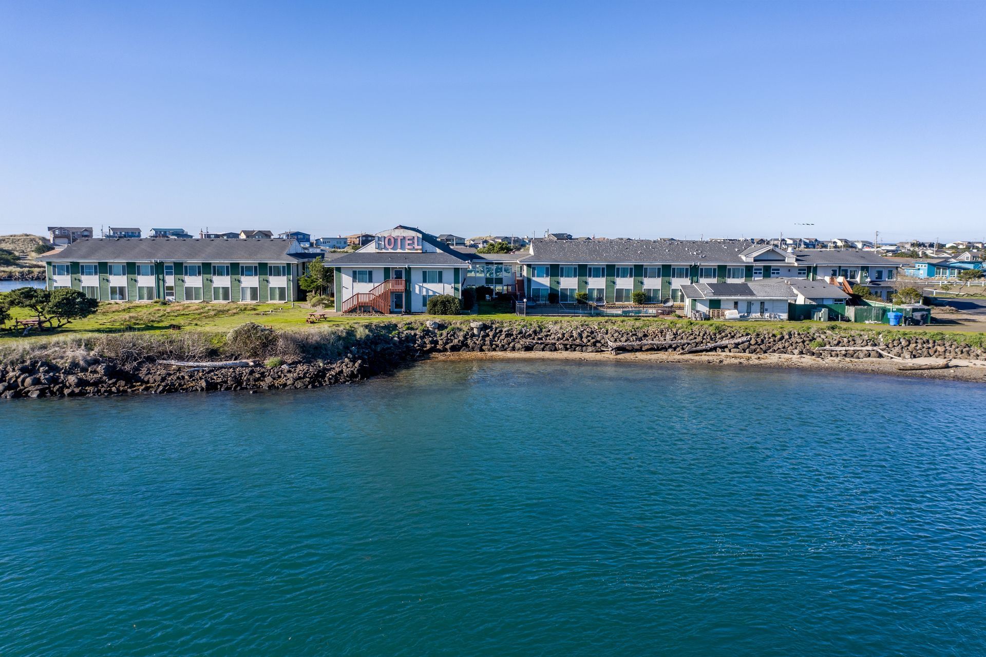 Waterfront resort buildings with green trim, next to blue water and a rocky shore, under a clear sky.