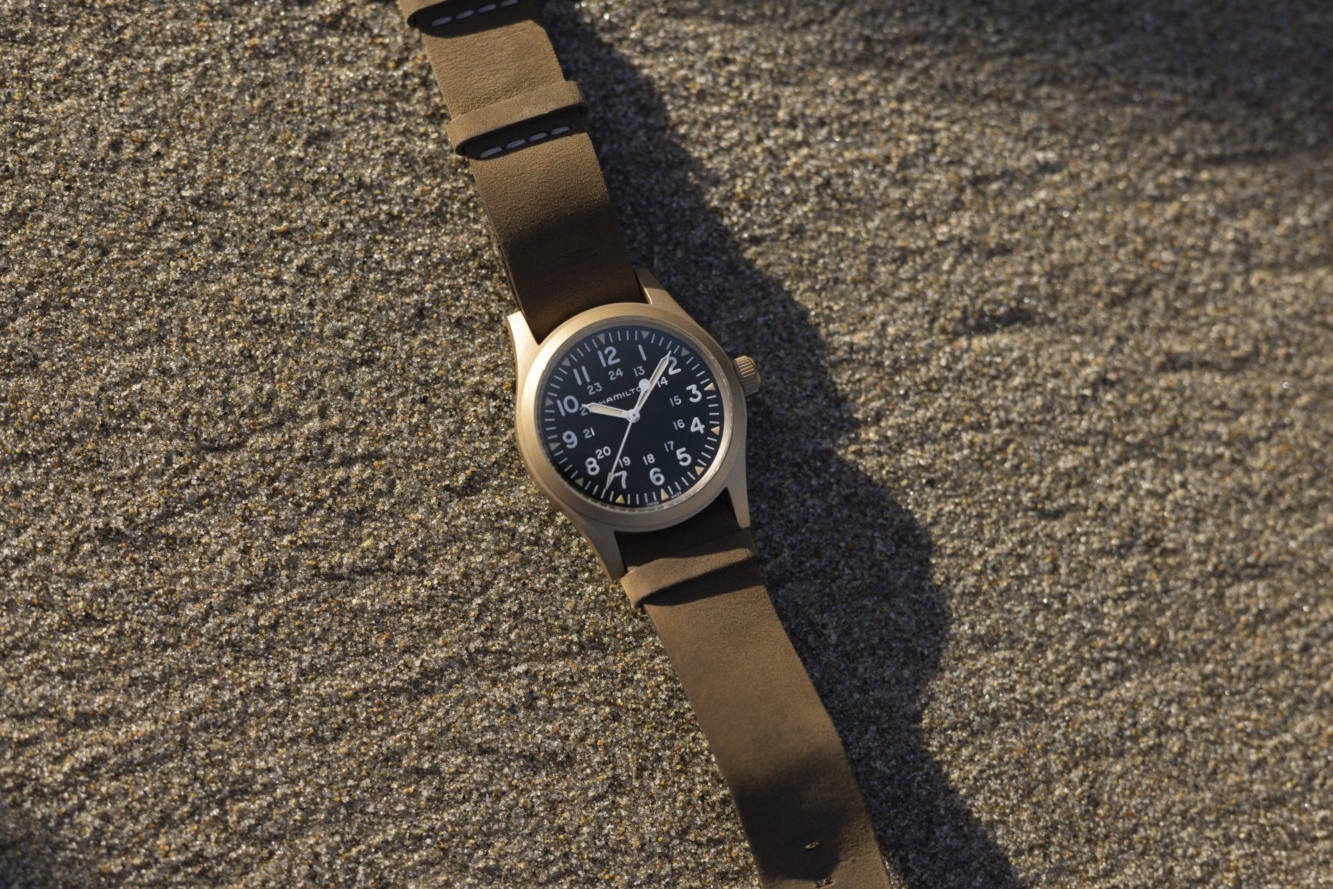 Orologio da polso nero con cinturino marrone, appoggiato su sabbia ruvida alla luce del sole.