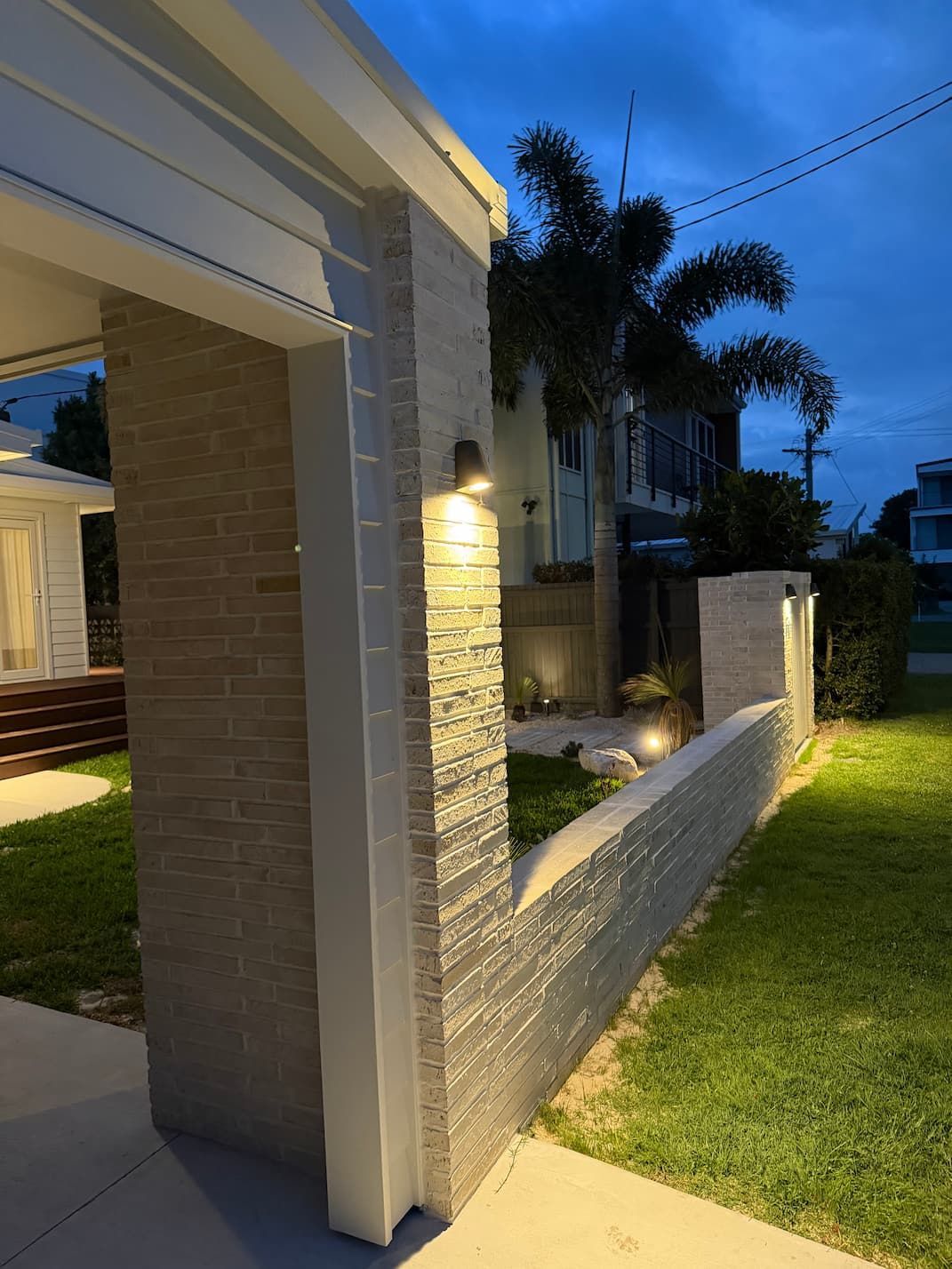 Brick and stucco pillars with warm lighting frame a pathway and a building at dusk.
