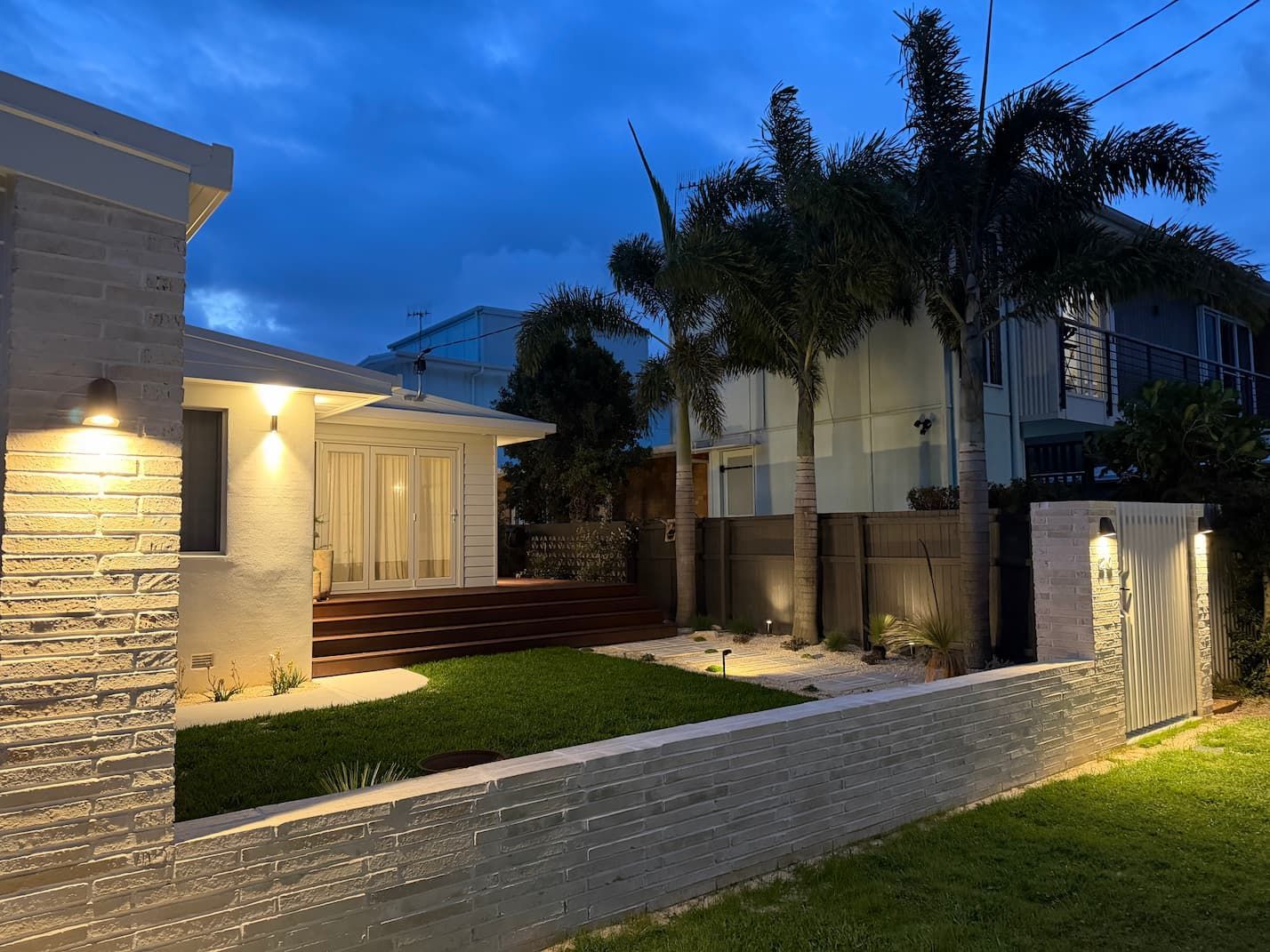 Night exterior view of a house with white walls, a small lawn, and palm trees; illuminated by lights.