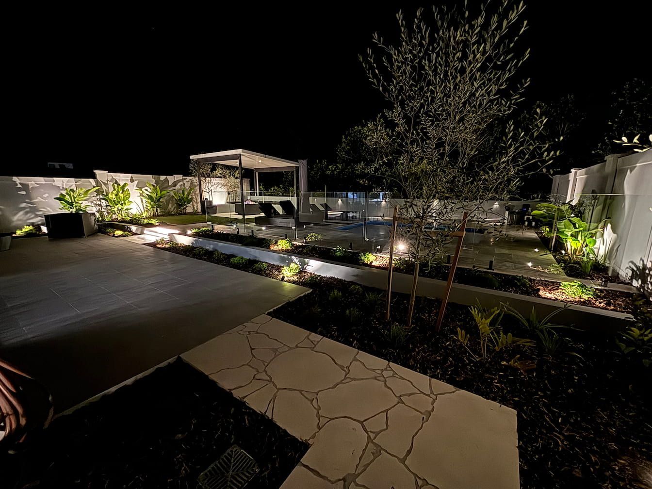 Night view of a lit backyard patio with a cabana, landscaping, and a dark sky.