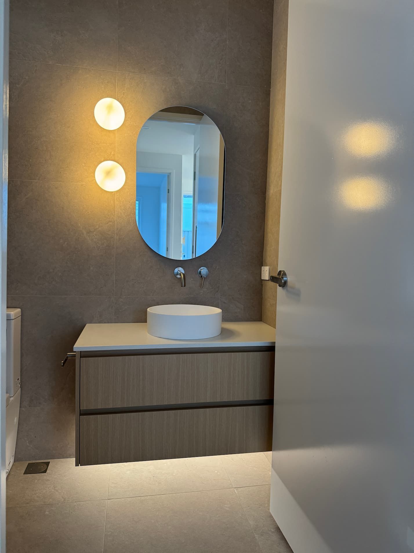 Bathroom with oval mirror, wood vanity, and round sink. Two sconces flank the mirror.