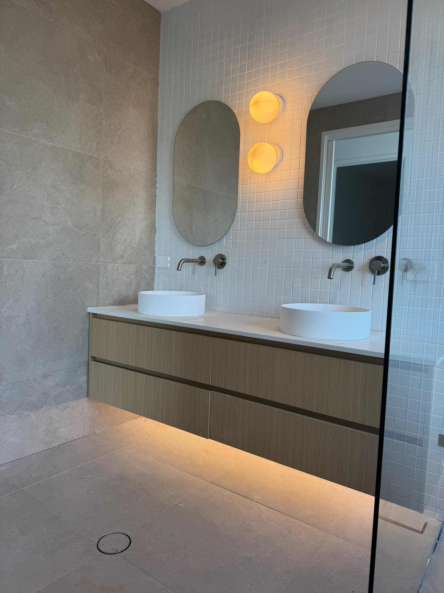 Modern bathroom with floating wooden vanity, two sinks, oval mirrors, and wall-mounted faucets.