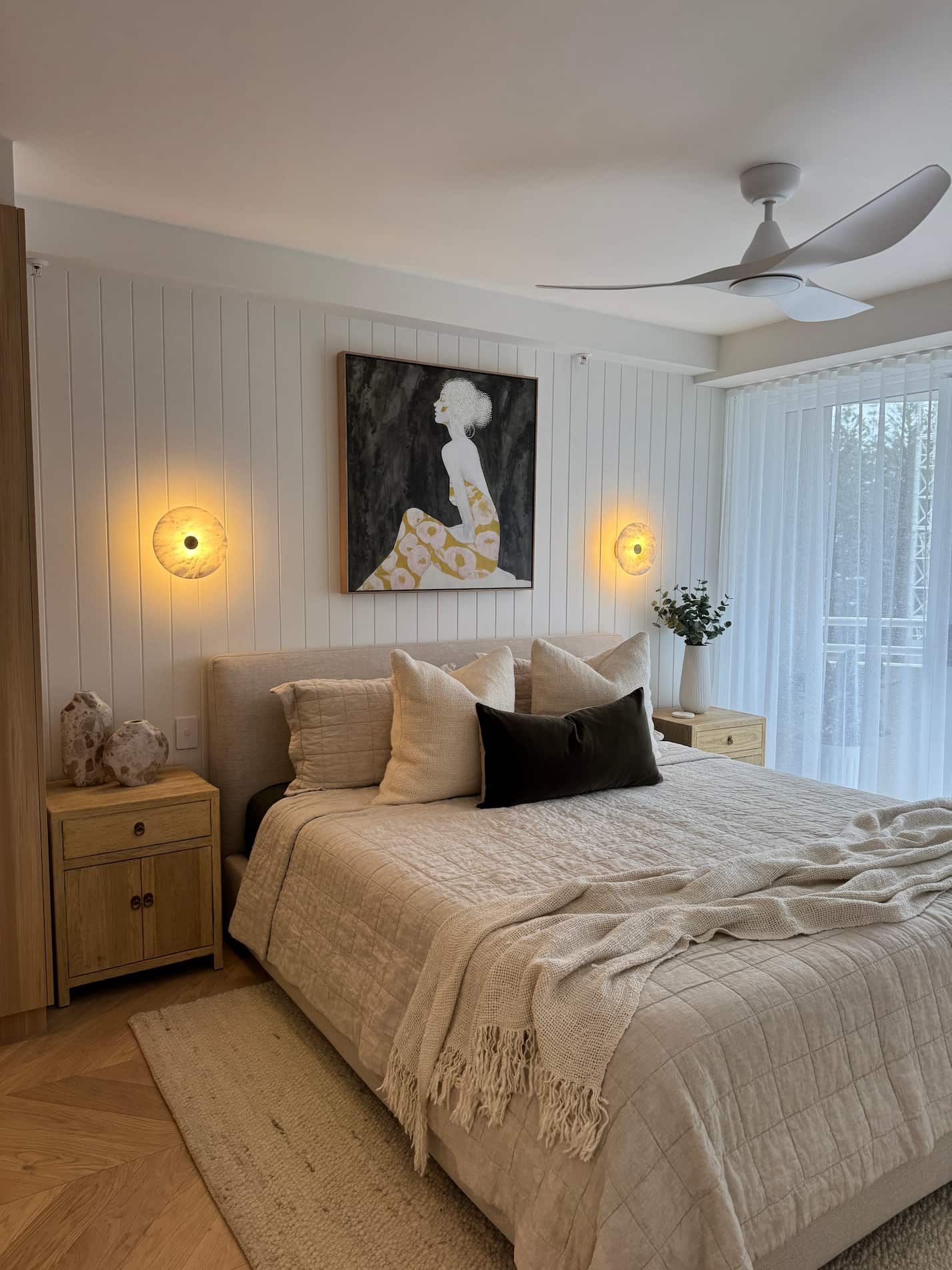 Bedroom with white paneling, art, bed, nightstands, and a ceiling fan.
