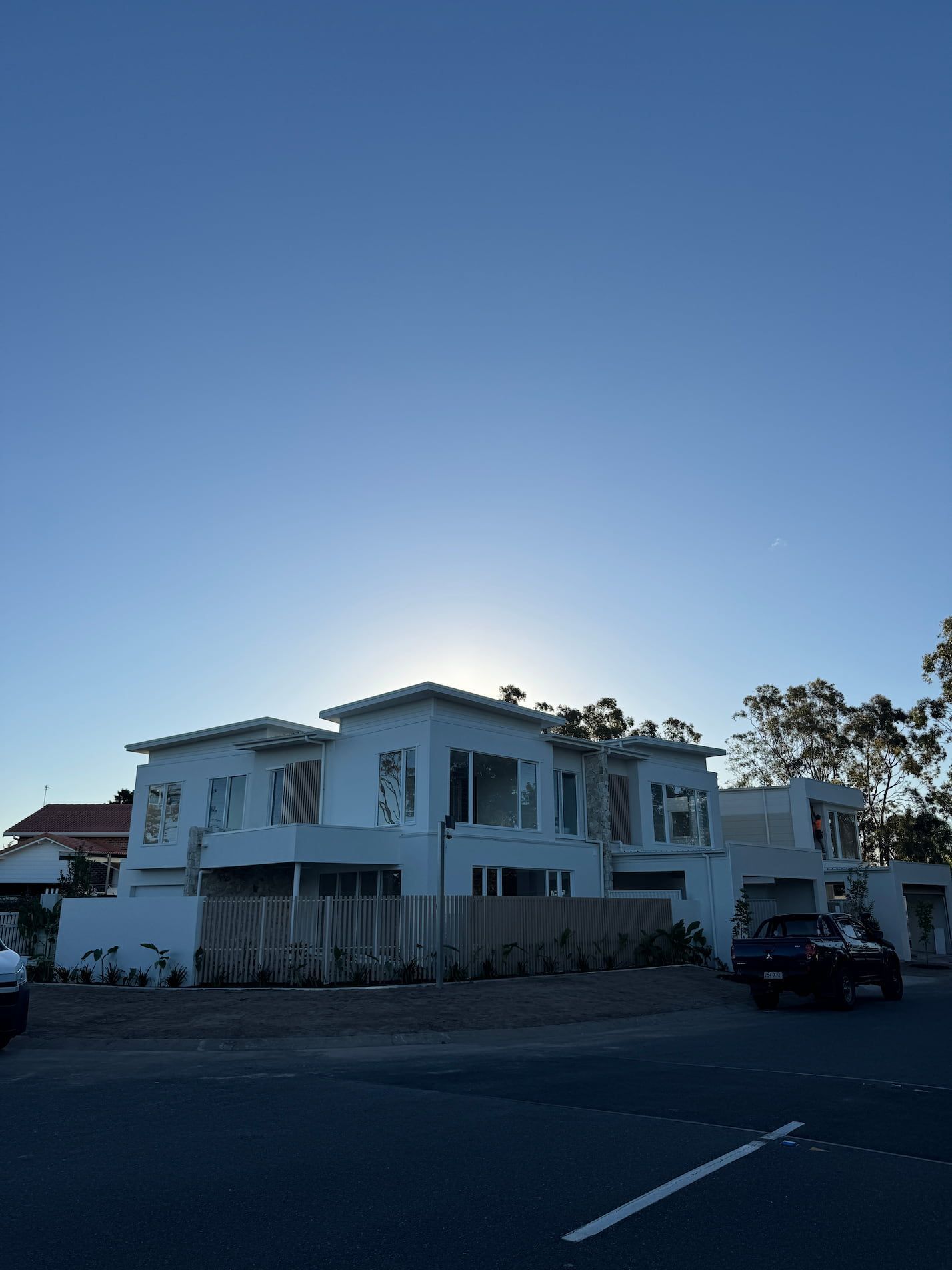 White house under construction with a blue sky, sun peeking over roof, and a car parked on the street.