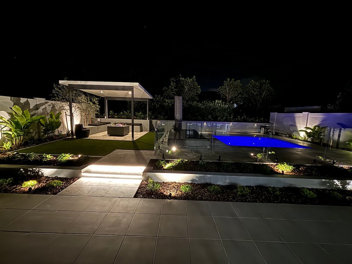 Night view of a backyard with a lit pool, patio, gazebo, and landscaping.