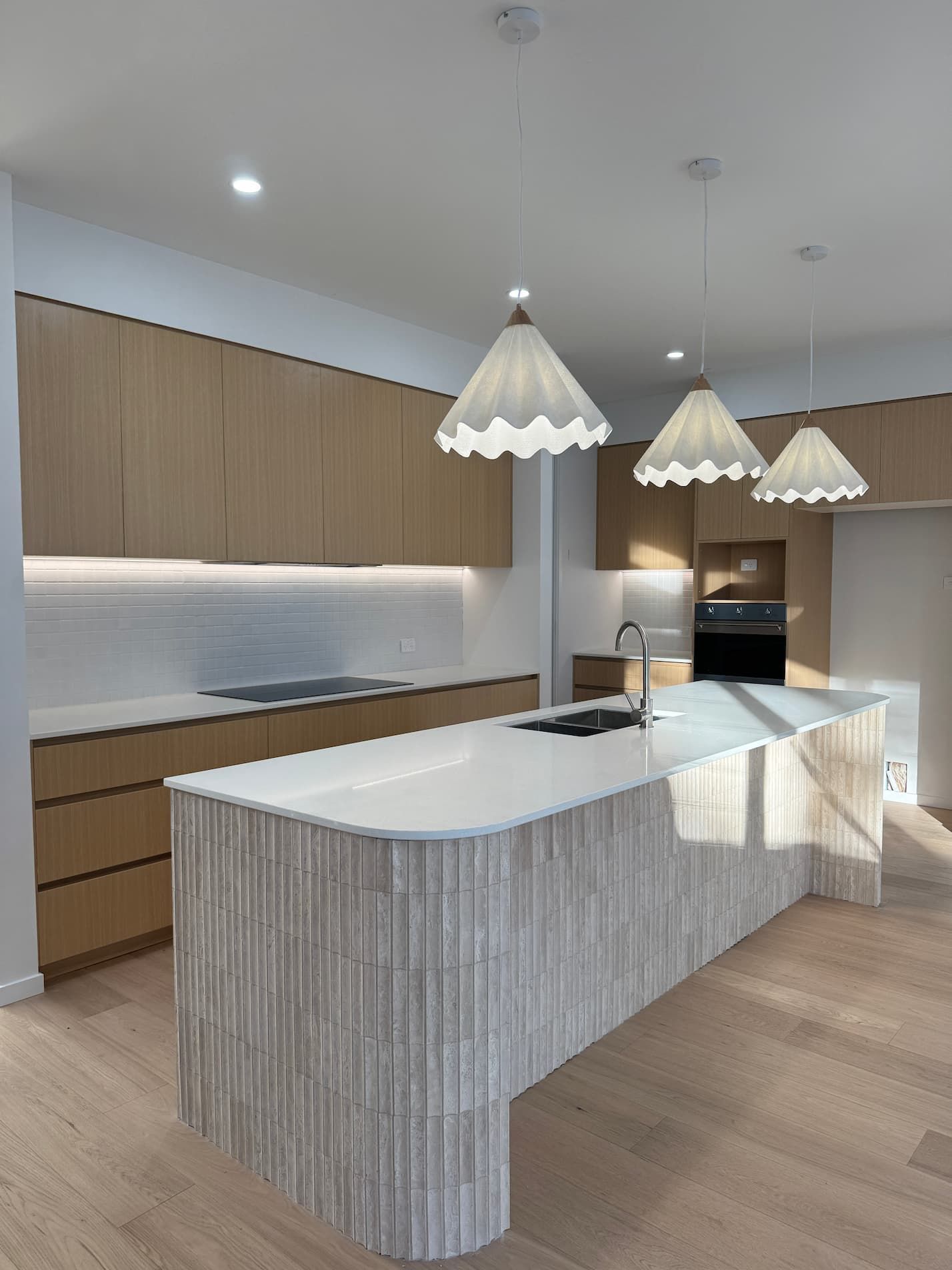 Modern kitchen with light wood cabinets, white countertops, and three pendant lights.