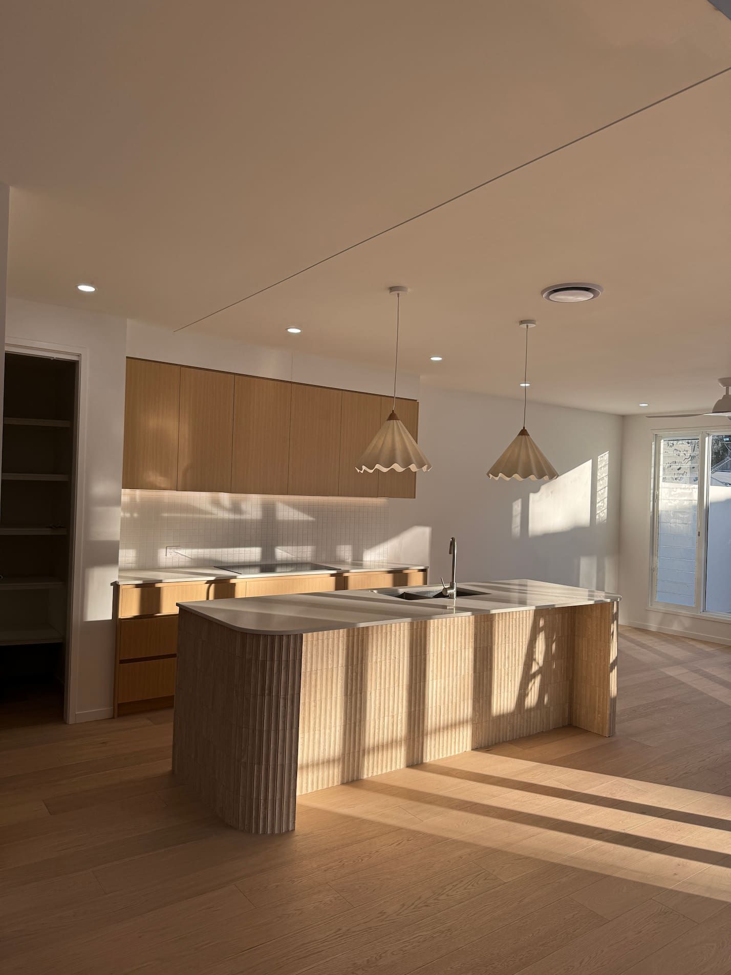 Modern kitchen with a light wood island and cabinets. Two pendant lights hang over the island. Sunlight streams in.