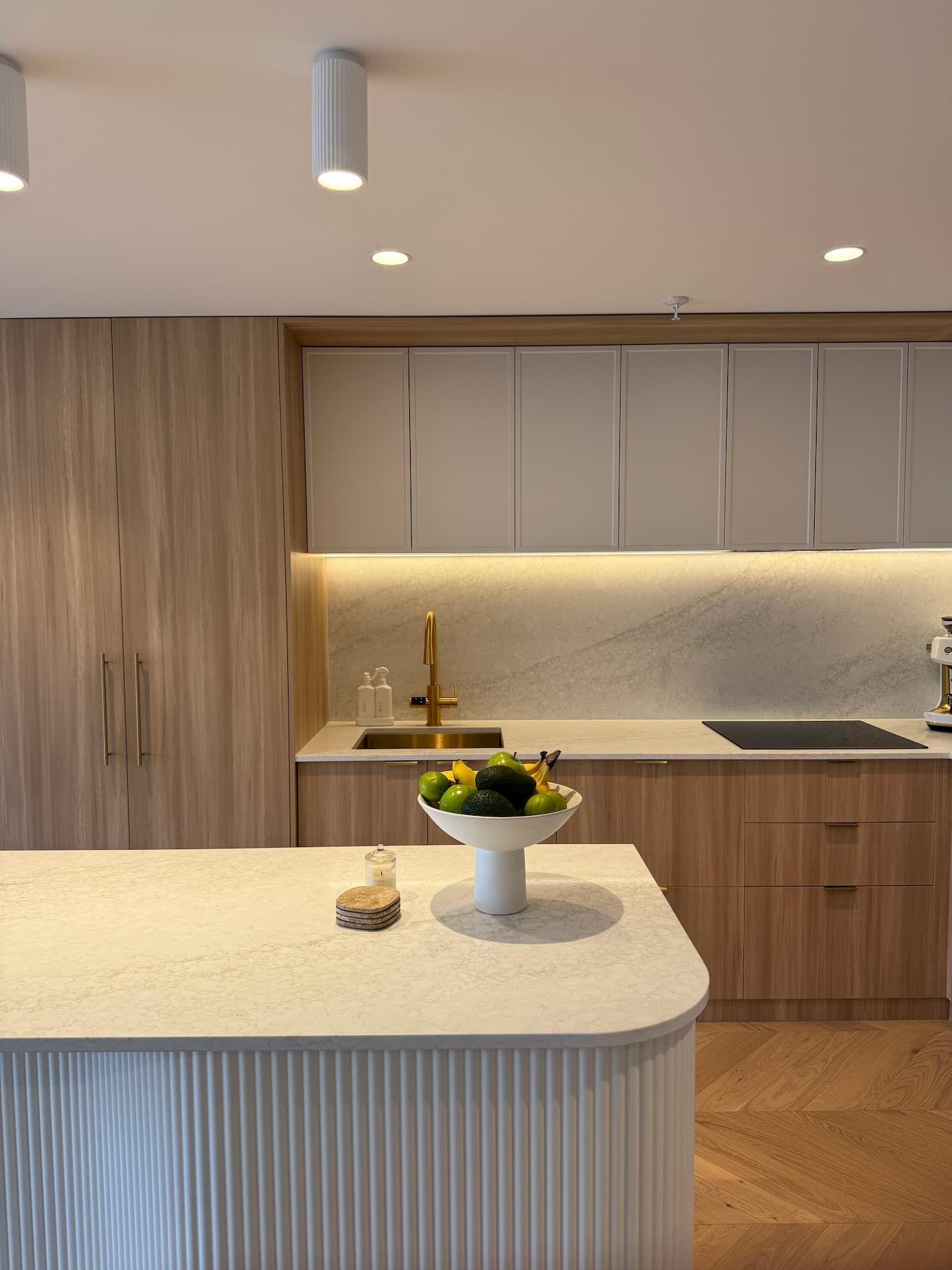 Modern kitchen with light wood cabinetry, white countertops, and an island. A fruit bowl sits on the island.