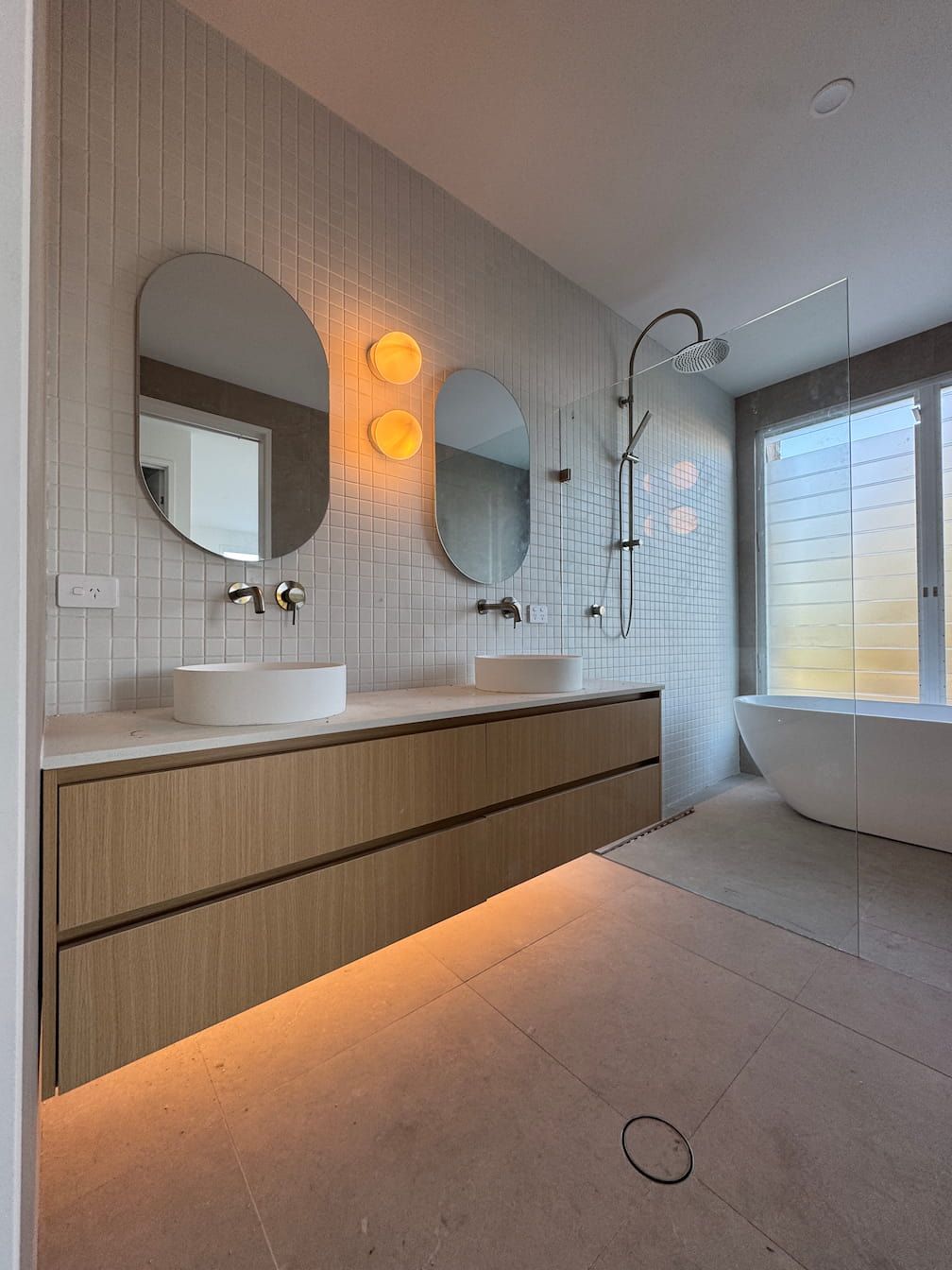 Bathroom with wood vanity, oval mirrors, and white sinks. Tile wall, glass shower, and freestanding tub.