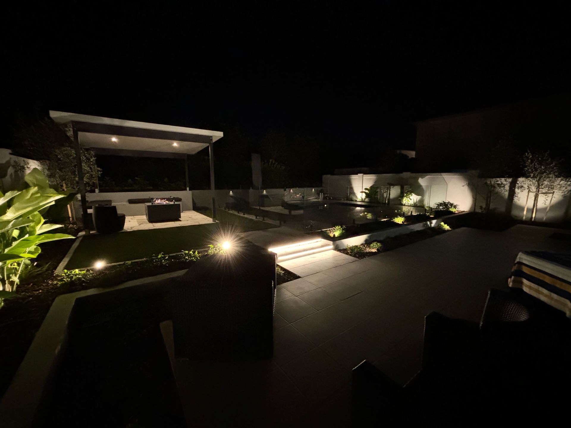 Outdoor nighttime scene with illuminated patio and steps, featuring a pergola and garden lighting.