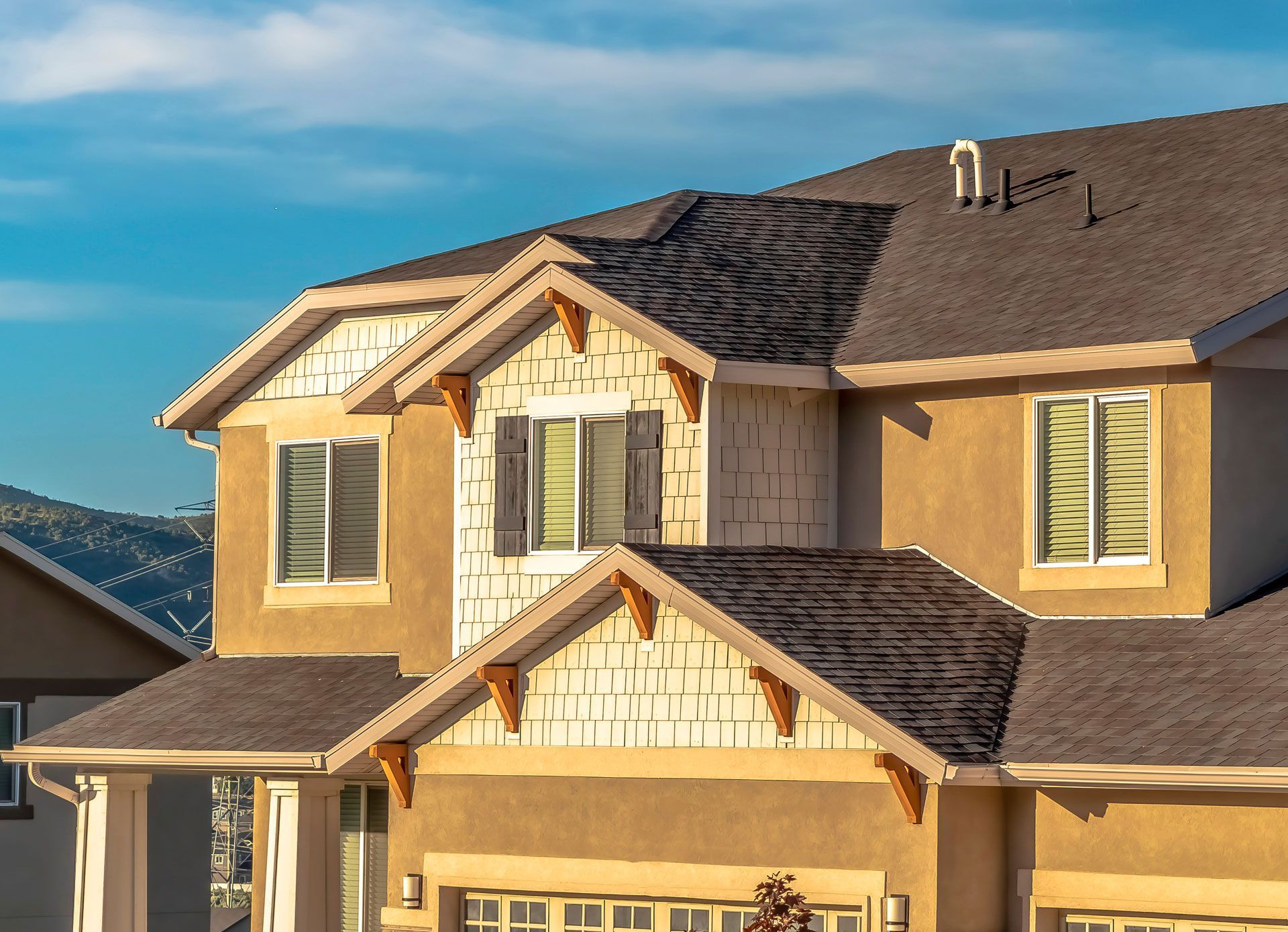 Two-story house with tan stucco walls, brown roof, and wooden accents against a blue sky.