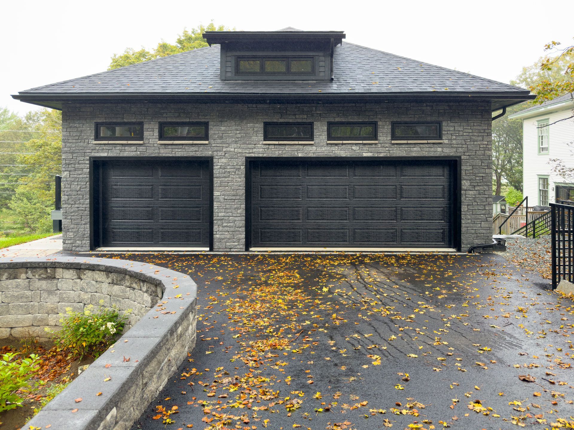 Stone garage with black doors and trim; a dark gray driveway with autumn leaves; a retaining wall on the left.