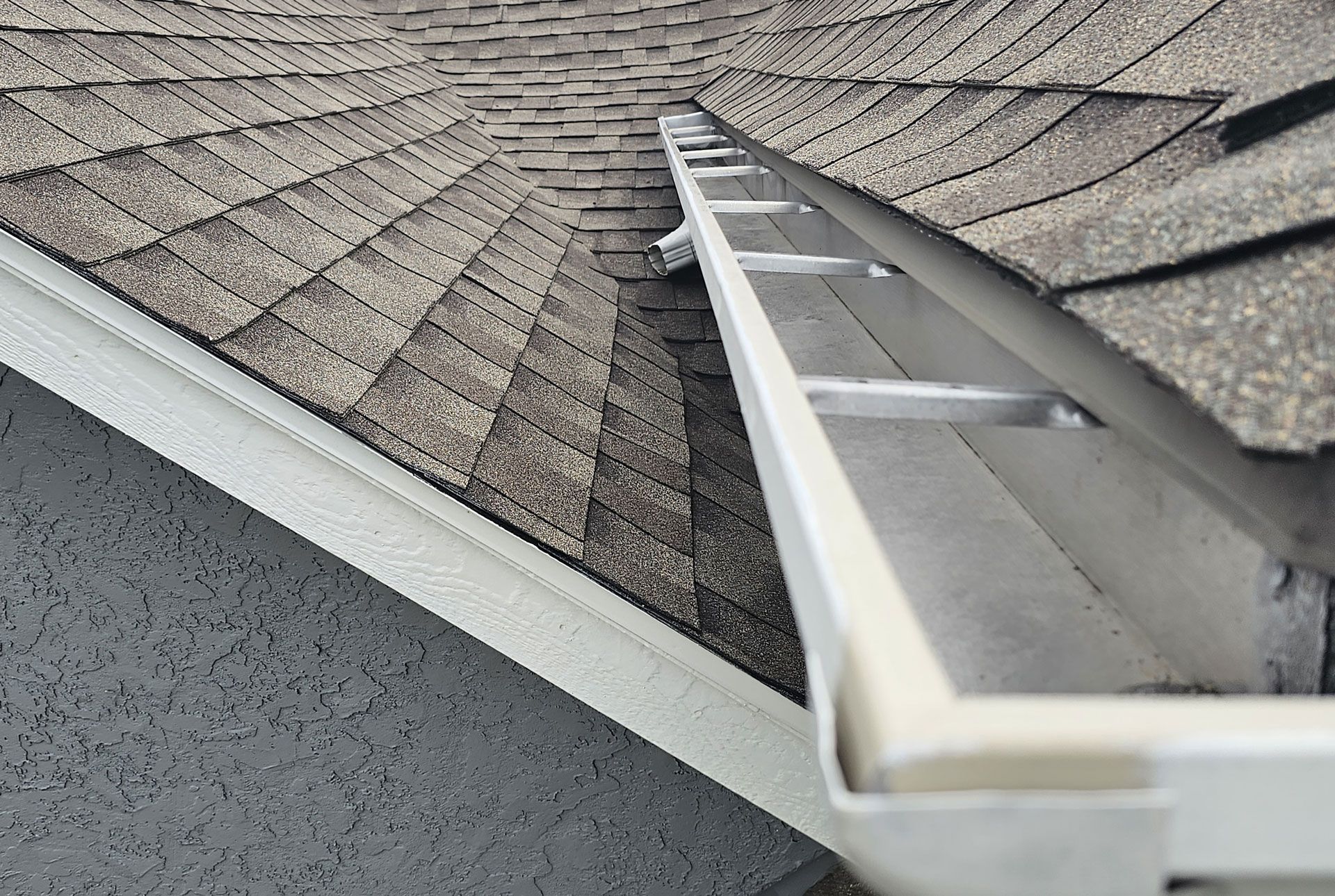 Gutter along a roof edge with asphalt shingles. The gutter is silver, the shingles are gray and the setting is outdoors.