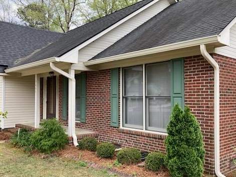 Small gray house with a wooden porch and a pathway, surrounded by a green lawn and flowering plants.