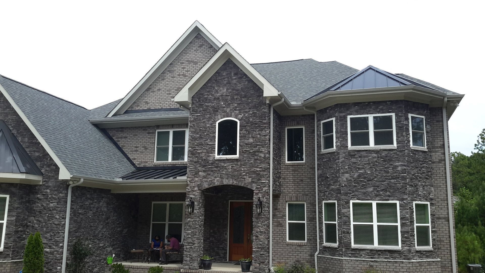 A two-story stone house with a gated driveway, surrounded by trees and green lawns under a blue sky.