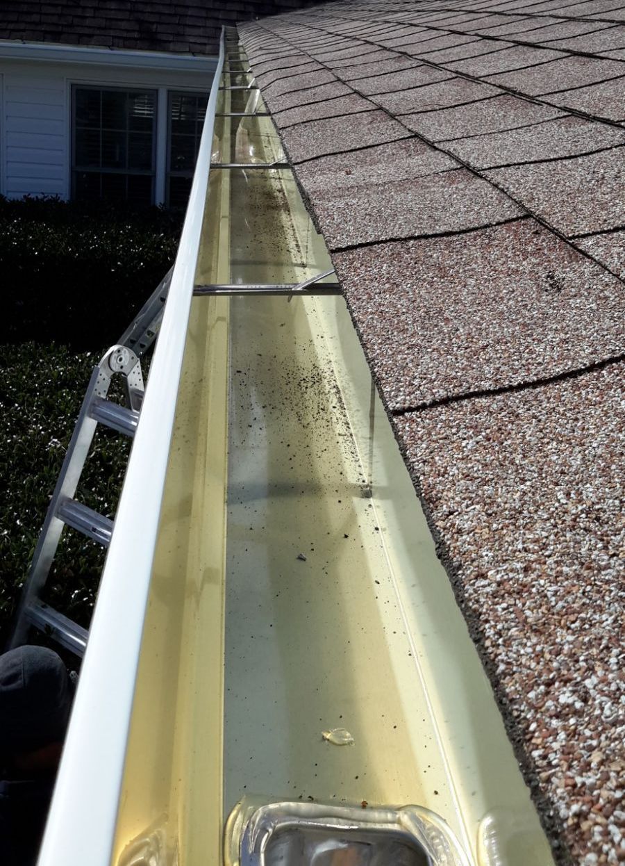Gutter guard installed on a roof, with black mesh over a dark gutter. Gray shingles and a white wall are also visible.