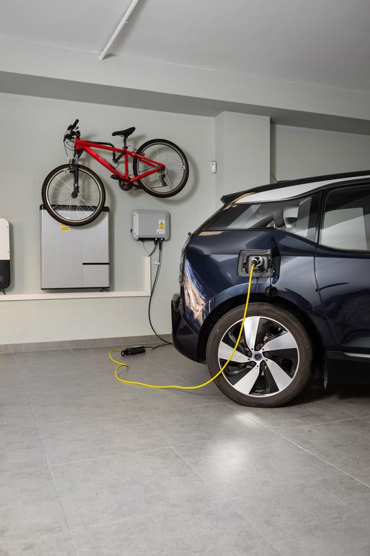Electric car charging in a garage; a yellow cable connects to the car. A bike hangs above a charging unit and energy storage system.